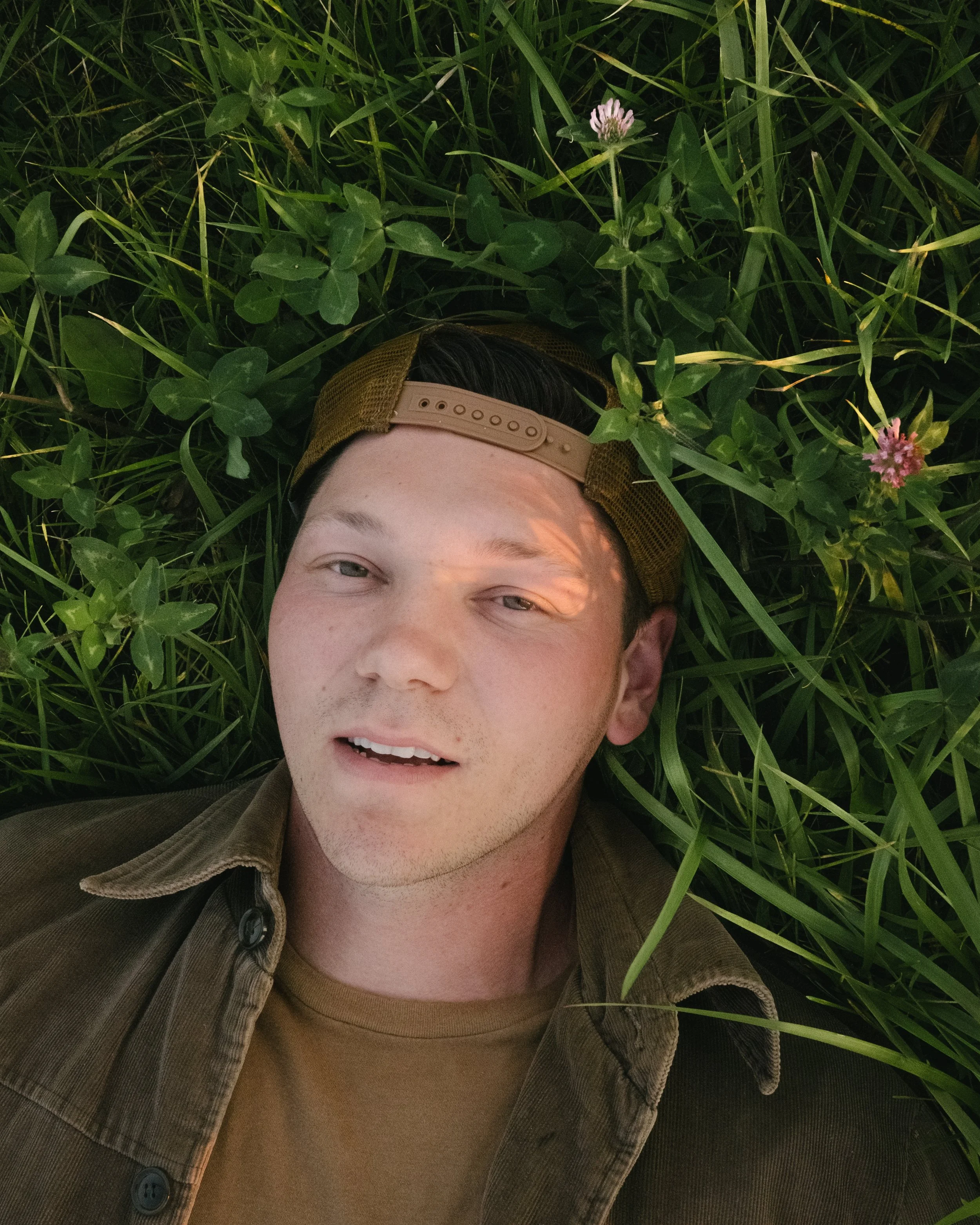 A young man with light skin and dark hair lying in green grass with pink flowers, wearing a brown shirt and a backward beige and brown cap.