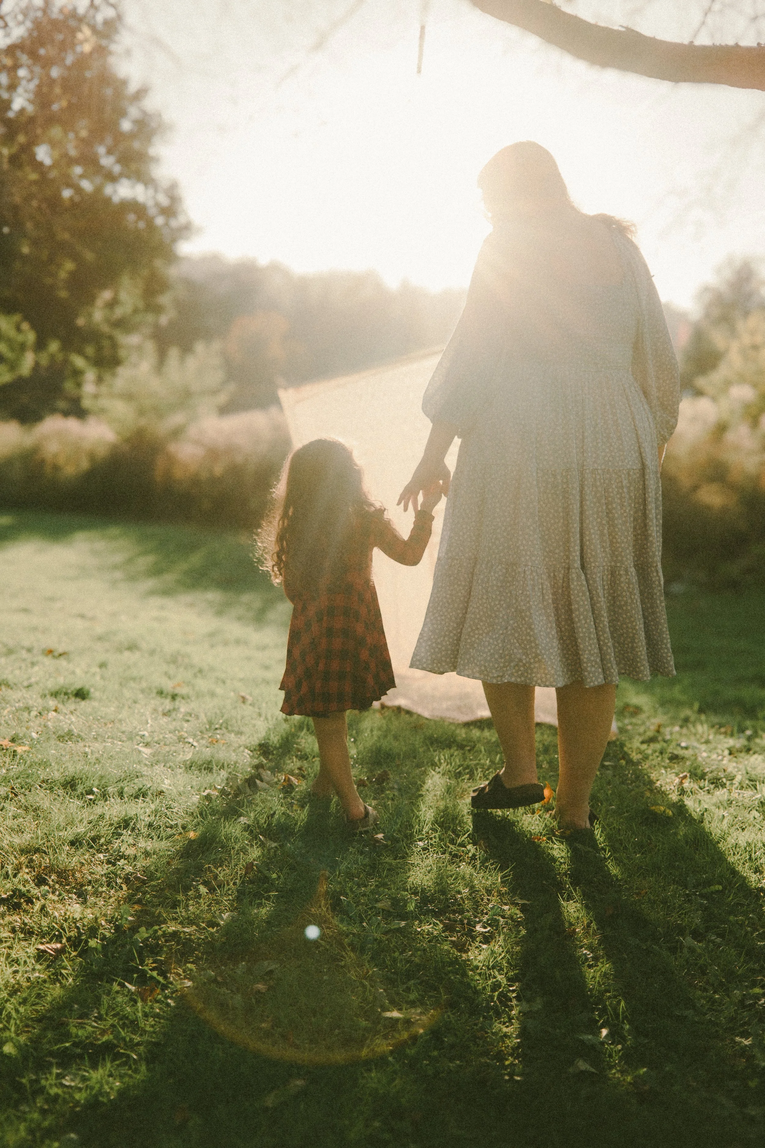 A woman and a young girl holding hands outdoors in sunlight, walking on grass near a teepee or tent, with trees and bushes in the background.