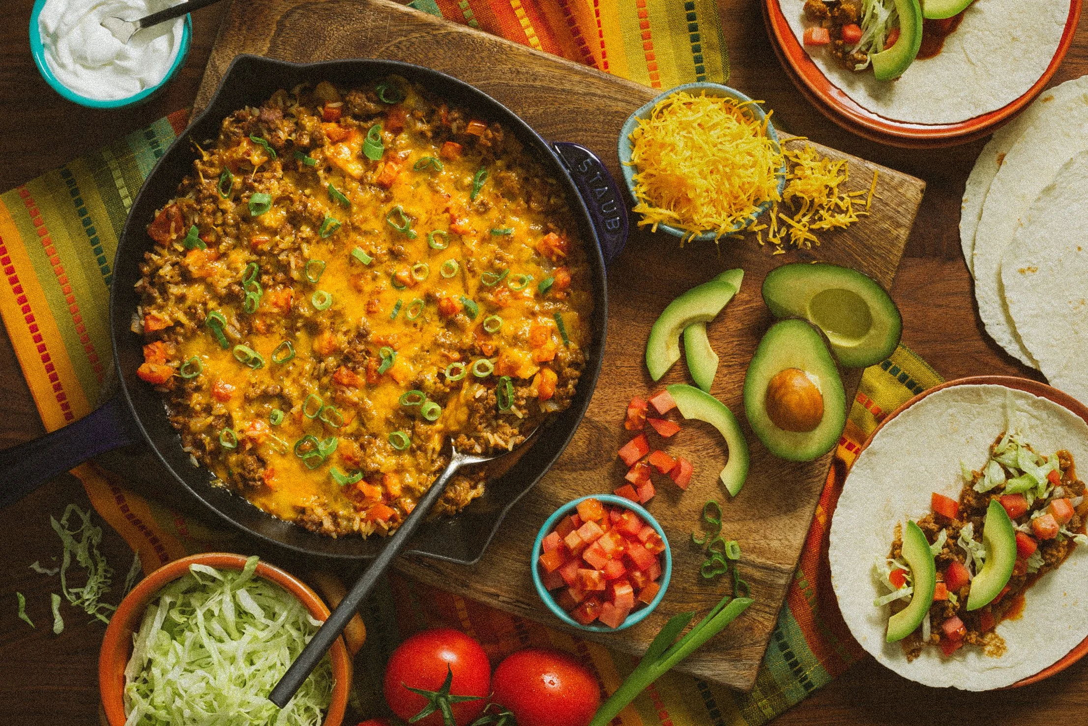 A spread of Mexican food featuring a skillet of cheesy beef and vegetable dip topped with green onions, surrounded by small bowls of shredded cheese, diced tomatoes, sliced avocado, shredded lettuce, and pico de gallo on a wooden table with tortillas