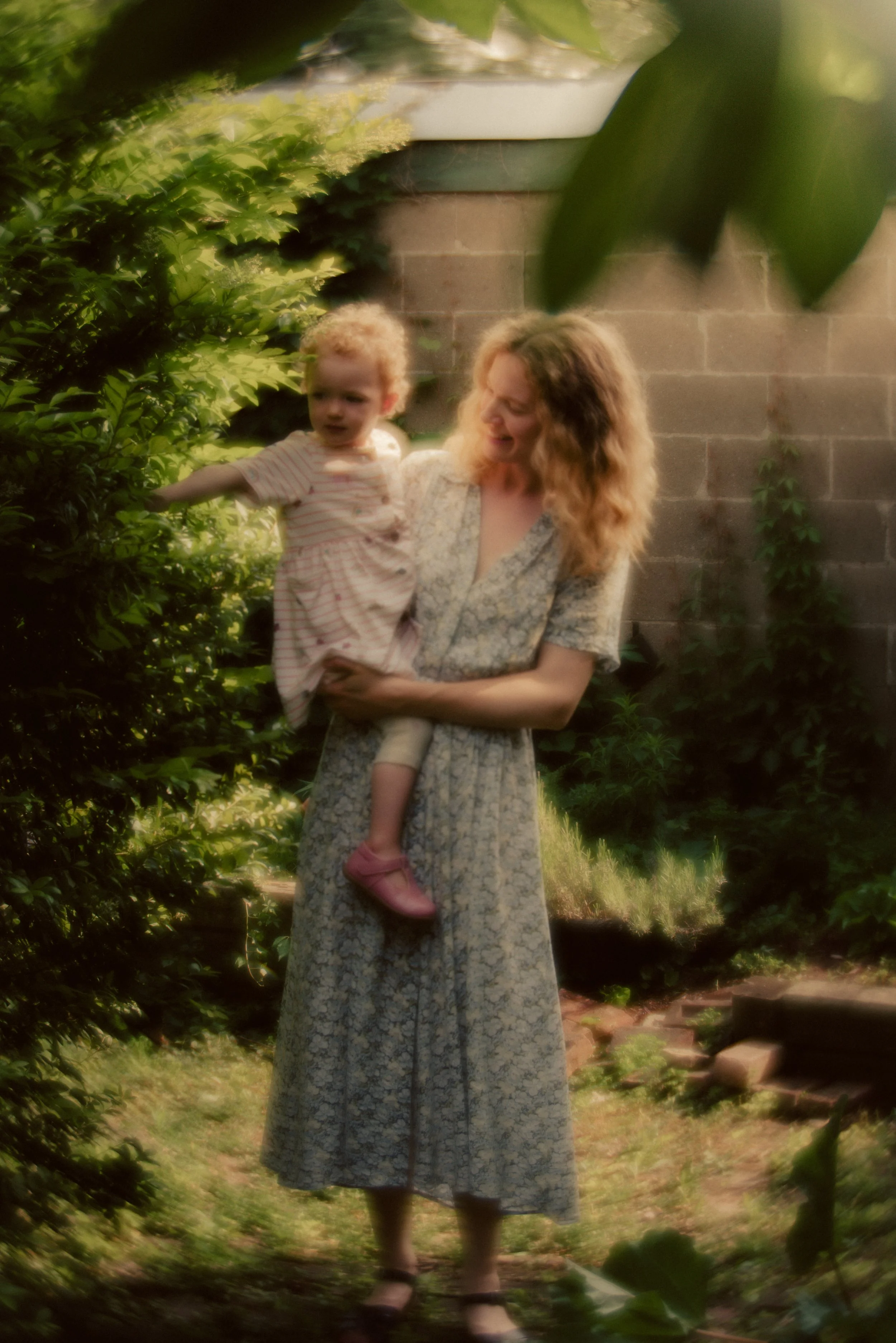 Outdoor portrait of a mother and young daughter with curly hair in a lush garden, with green foliage and a brick wall, capturing a tender moment
