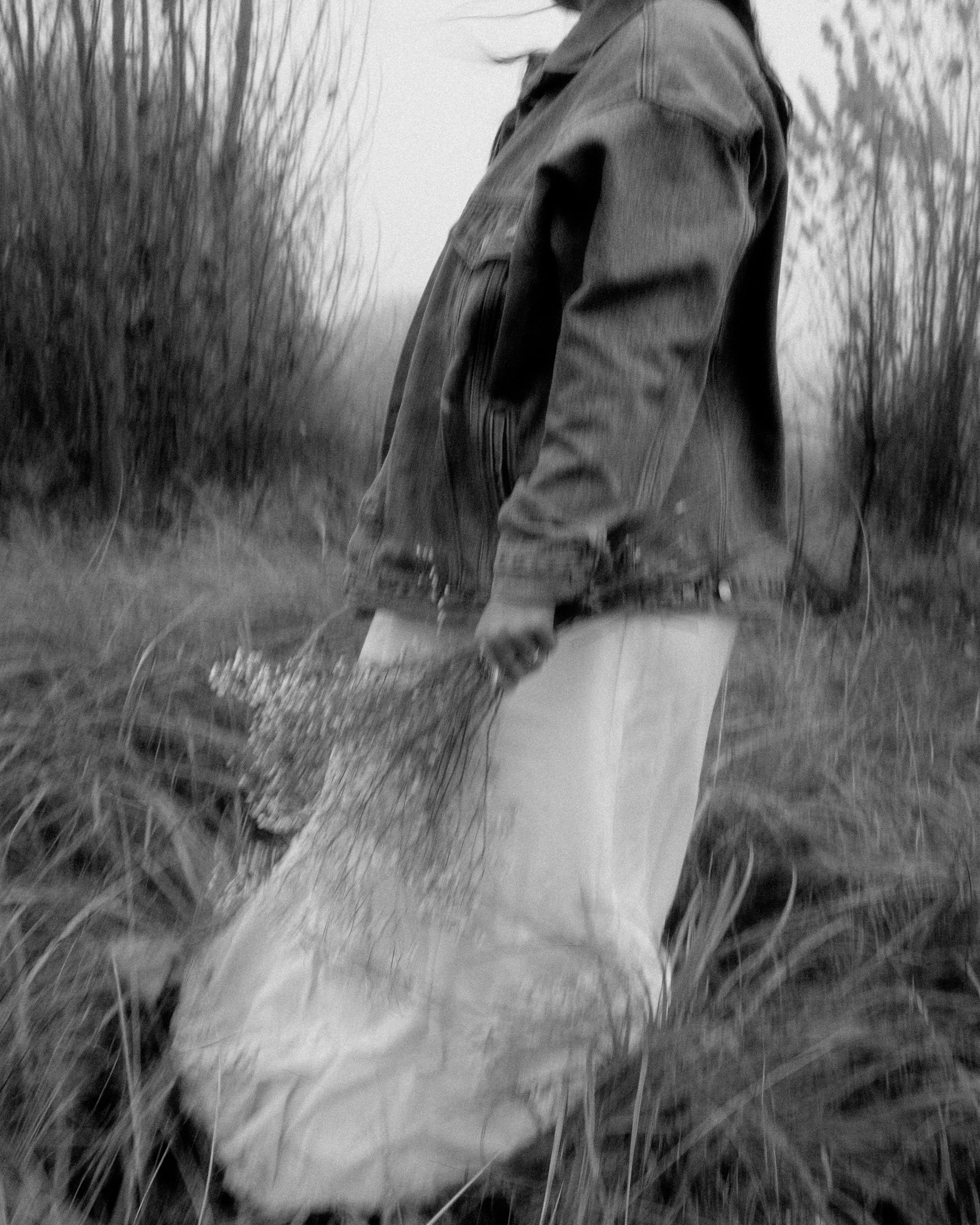 Person wearing a denim jacket and light-colored pants holding a net or bag in a field with tall grass and trees in the background.