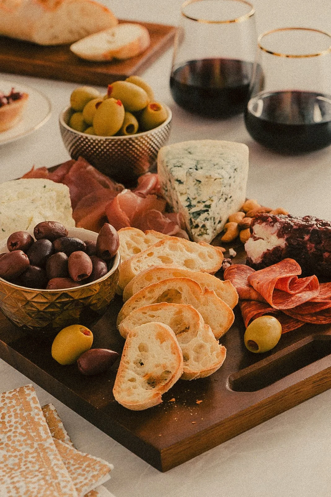 Charcuterie board with bread, olives, cheese, and meats, surrounded by wine glasses on a table.