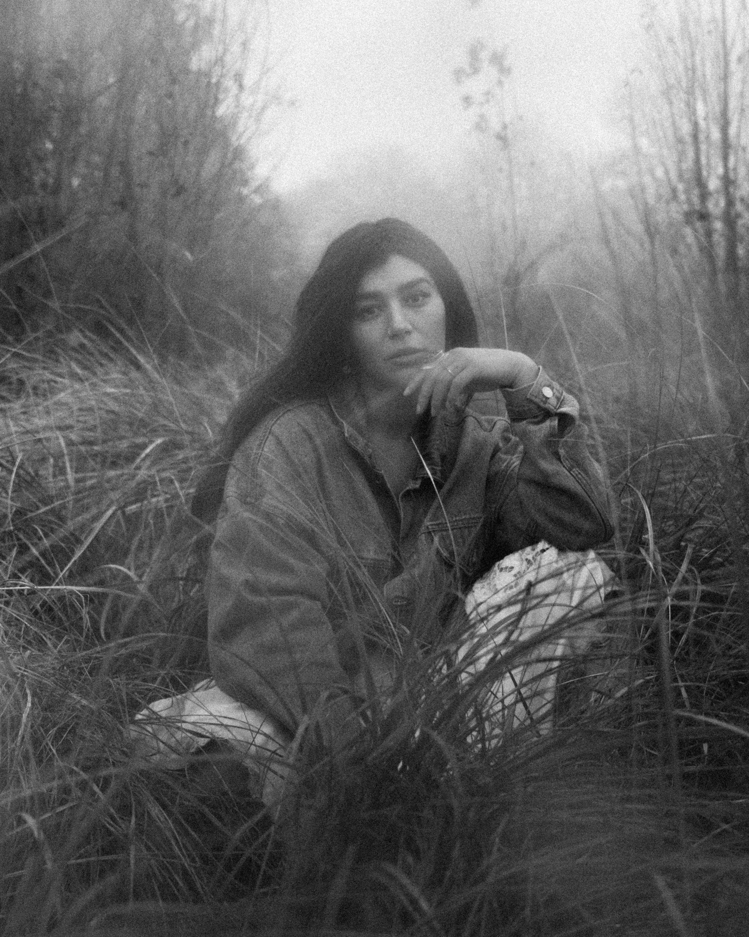 Black and white photo of a woman sitting among tall grass in an outdoor, wooded area, wearing a denim jacket, with long black hair and a contemplative expression.