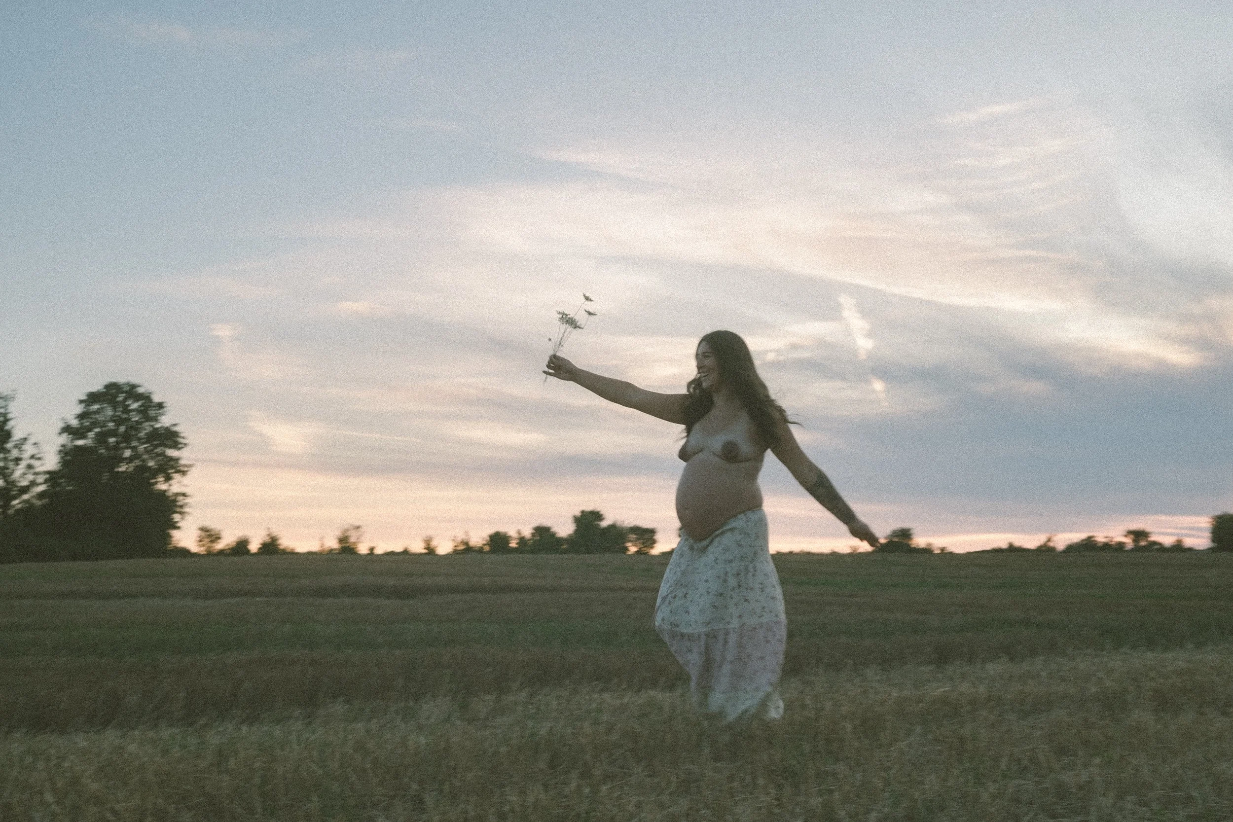 A pregnant woman in a flowing skirt standing outdoors at sunset, smiling and holding a small branch with leaves and flowers.