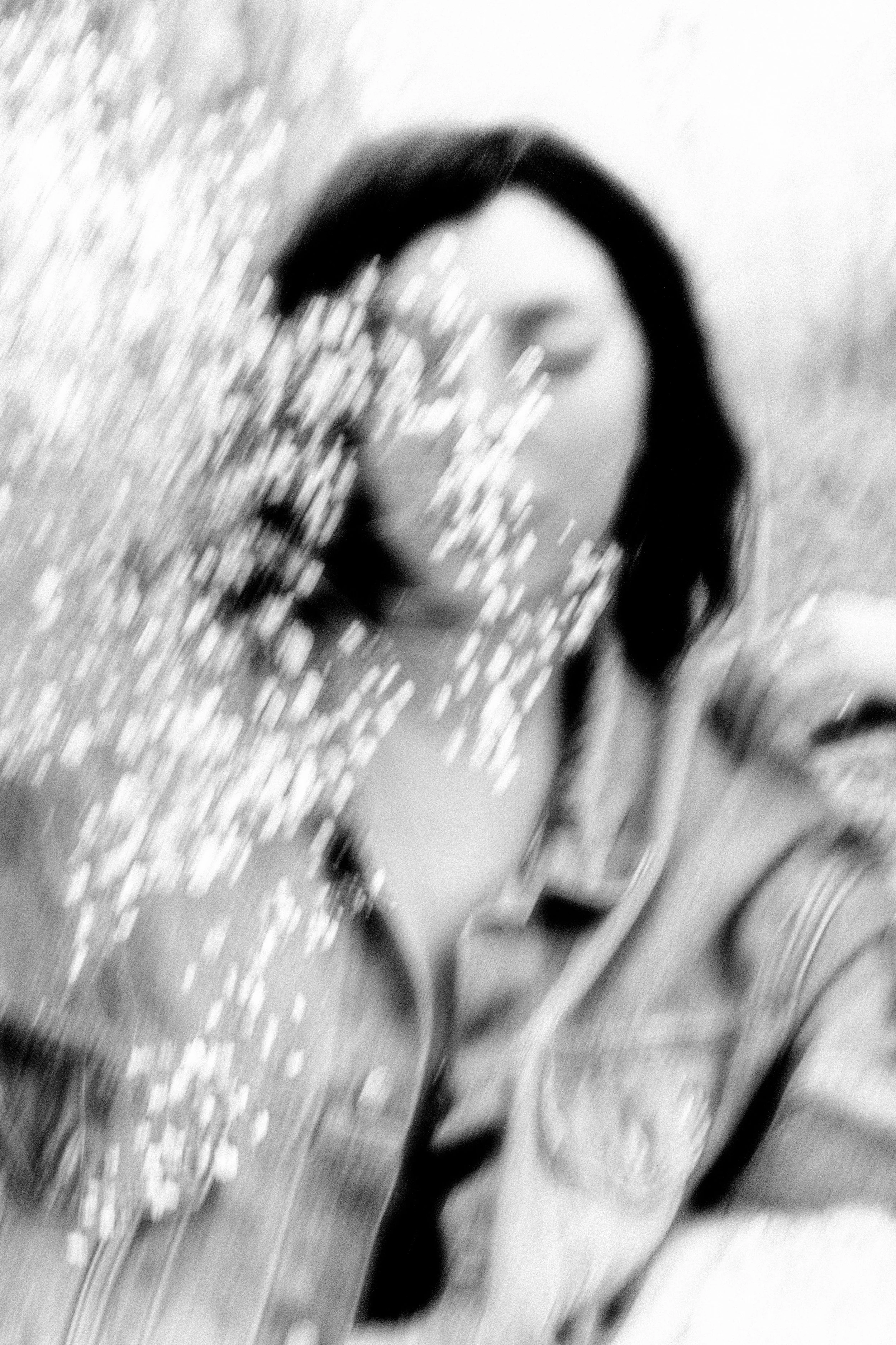 A black and white photo of a young woman with dark hair, wearing a denim jacket, holding and looking at a handful of small, shiny objects that could be beads or tiny crystals.