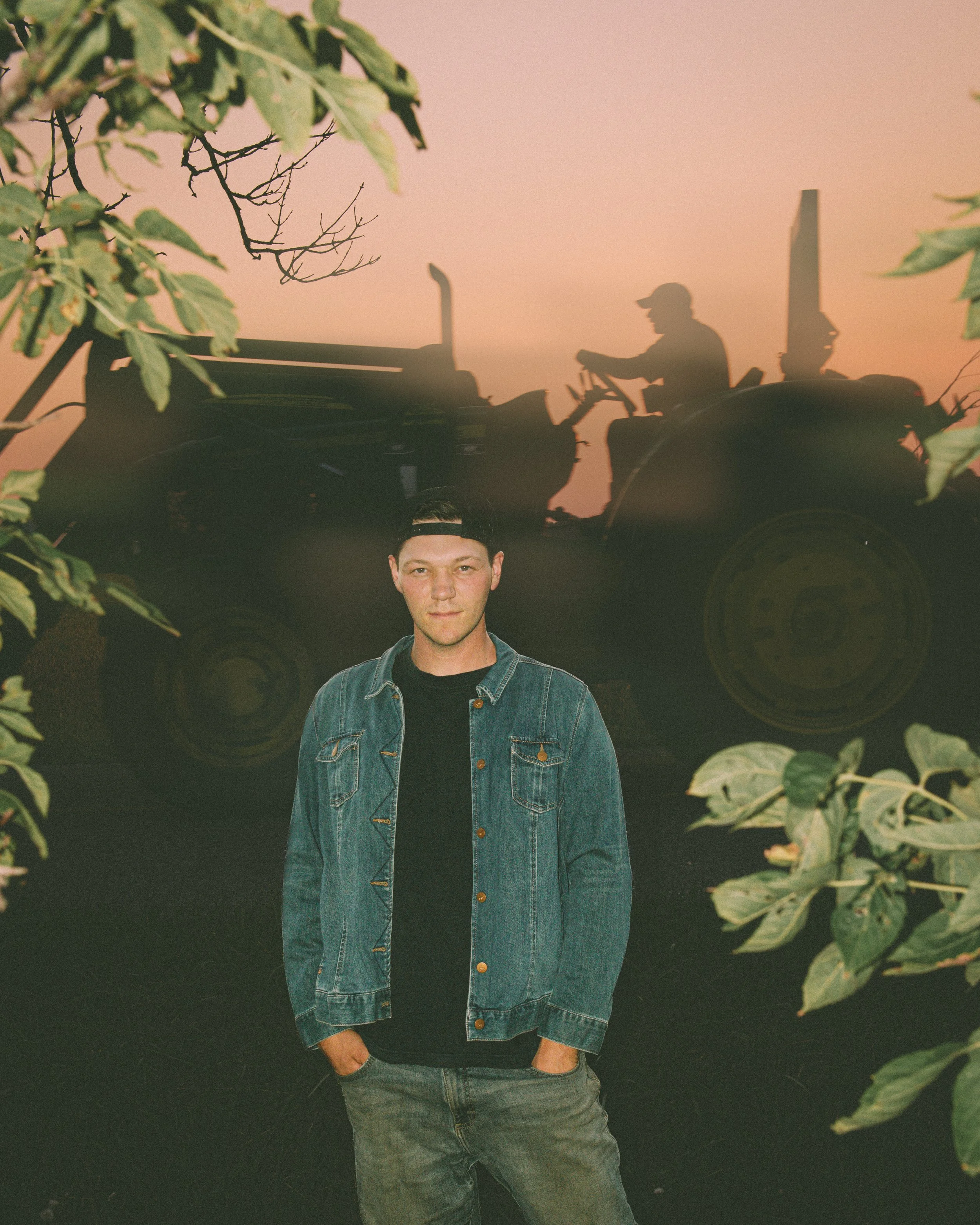 A young man stands outdoors during sunset, framed by green foliage, with a silhouette of a tractor and a person driving it in the background.