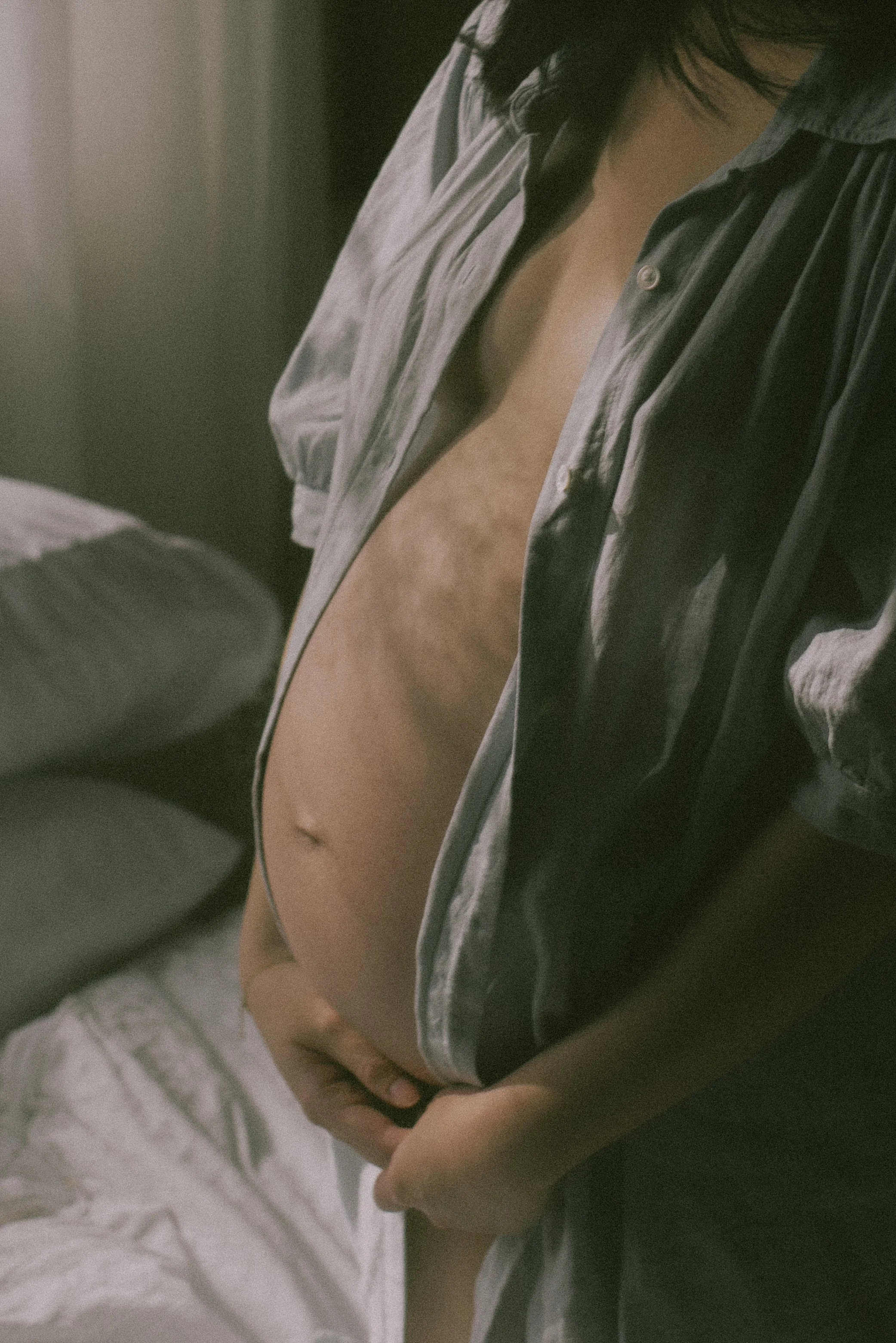 A person in a partially unbuttoned black shirt revealing a pregnant belly, standing in a dimly lit room.