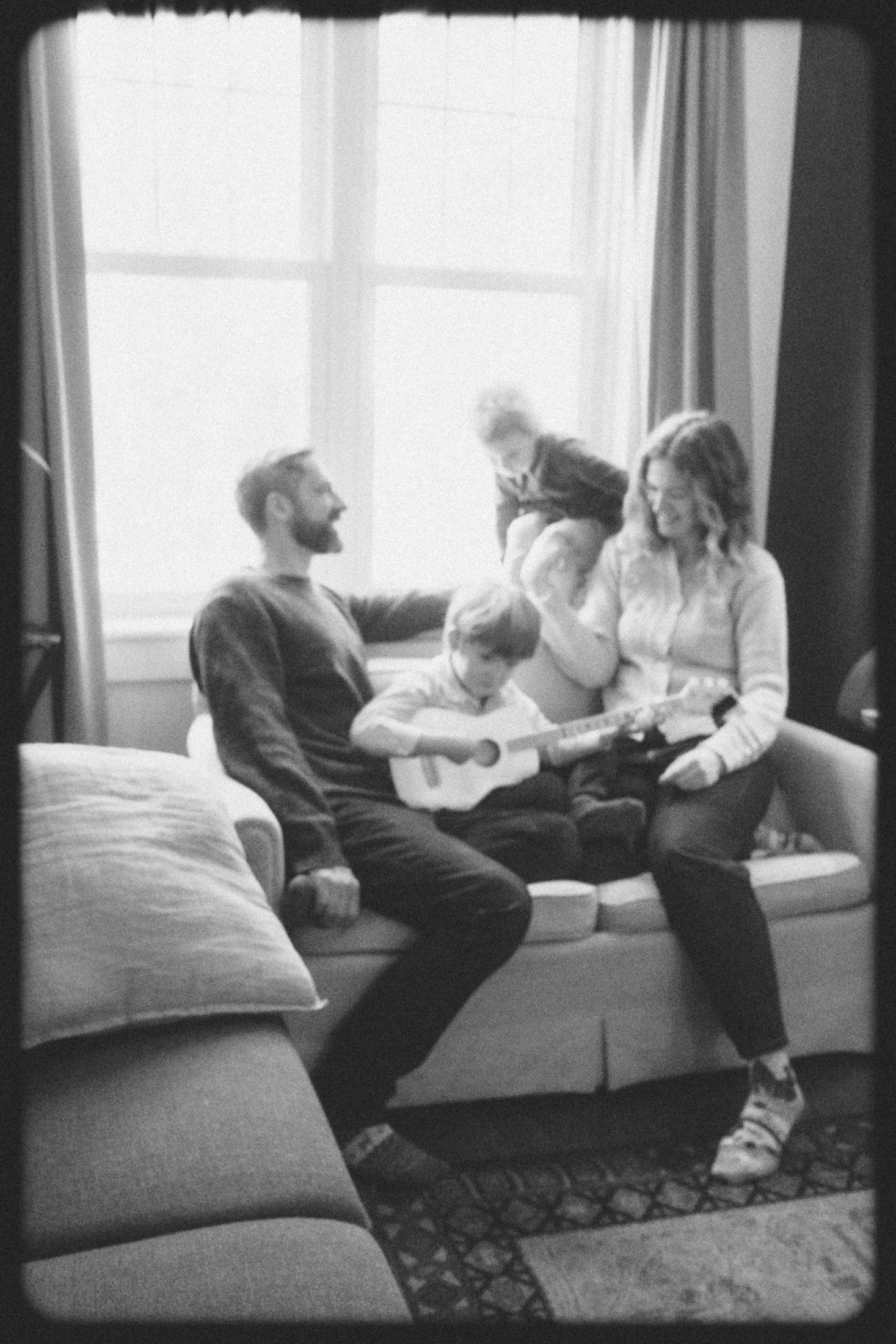 A family of four sitting on a couch in a brightly lit room. A child is jumping on from behind on the top of the couch, near a window with curtains. another child is between mom and dad playing a small guitar