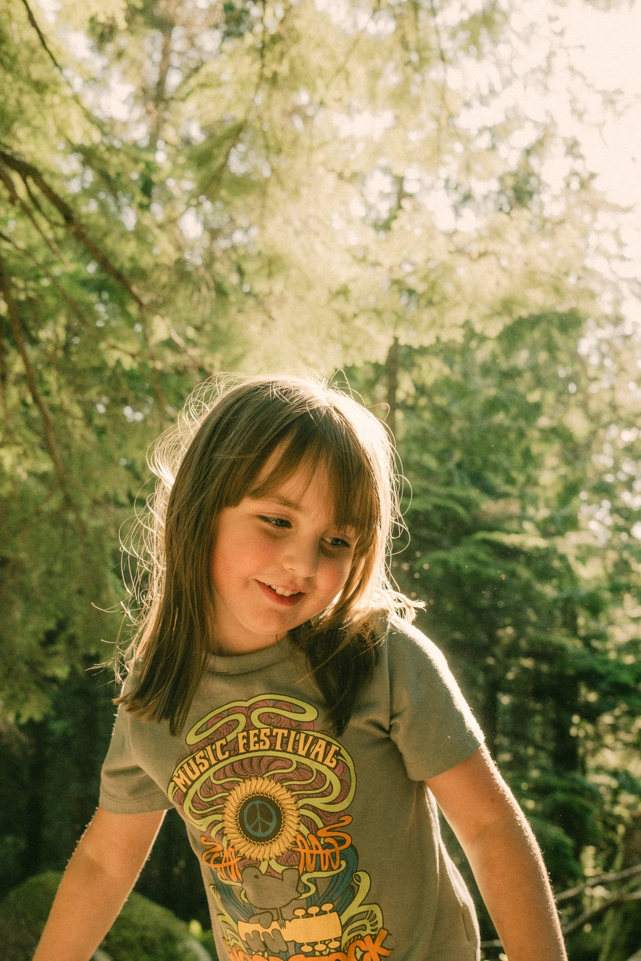 A young girl with shoulder-length brown hair standing outdoors in a forested area with sunlight filtering through the trees.
