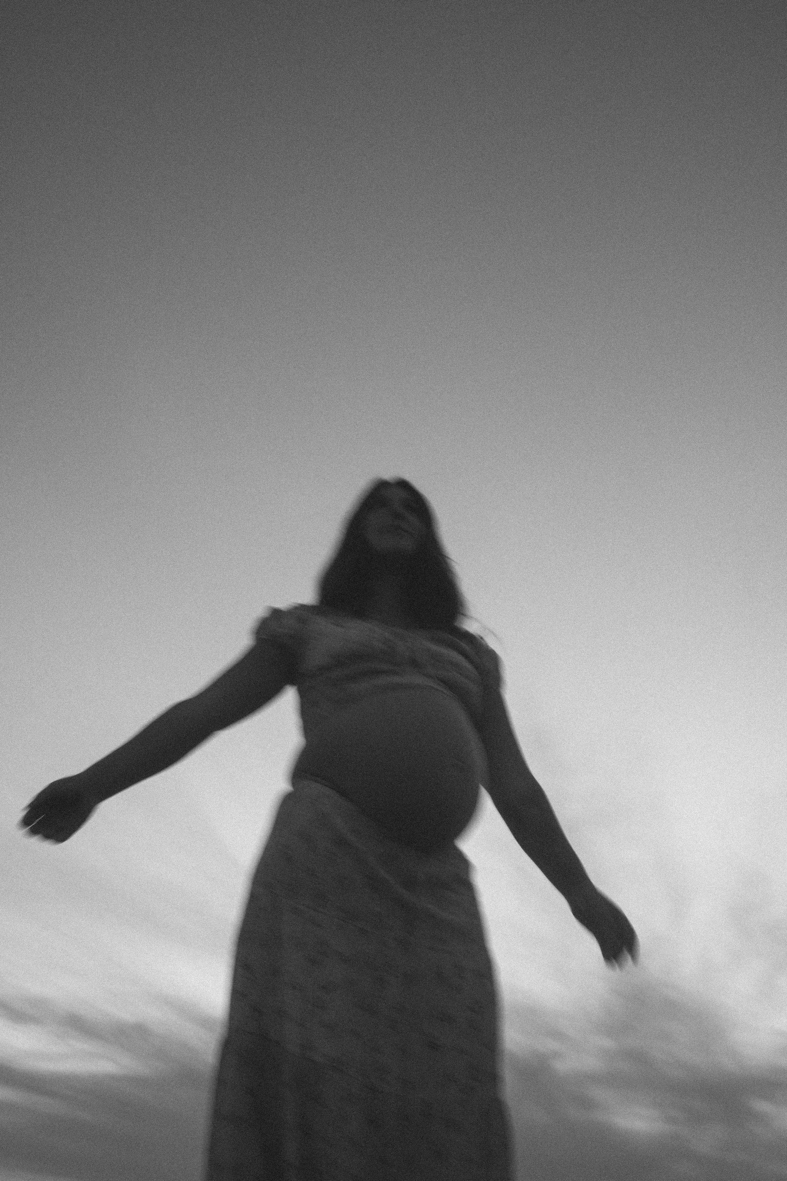 Black and white photo of a pregnant woman standing outdoors with arms outstretched, looking up, against a clear sky.