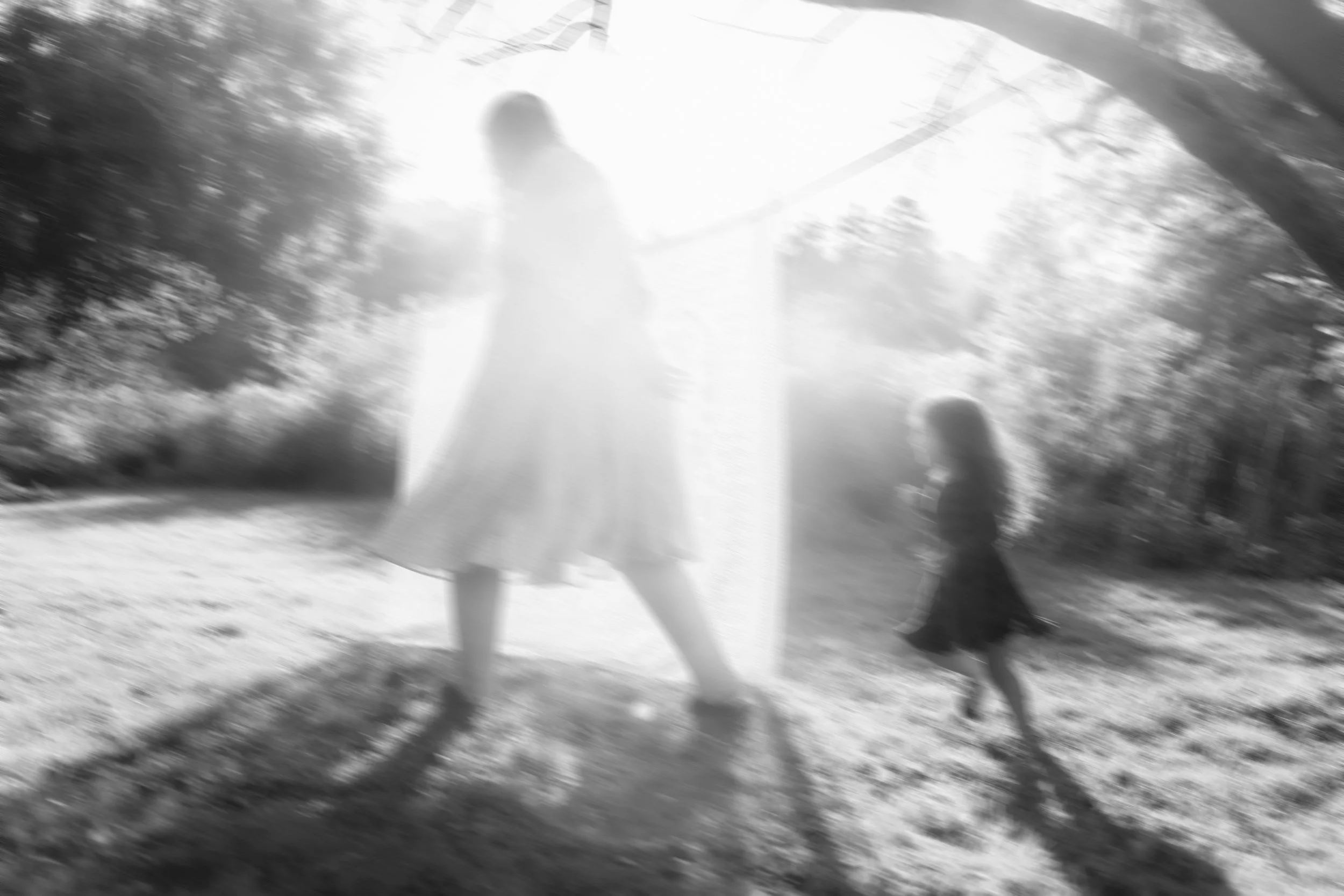 Blurred black-and-white photo of a woman and a child walking outdoors under a bridge or archway, with trees in the background.