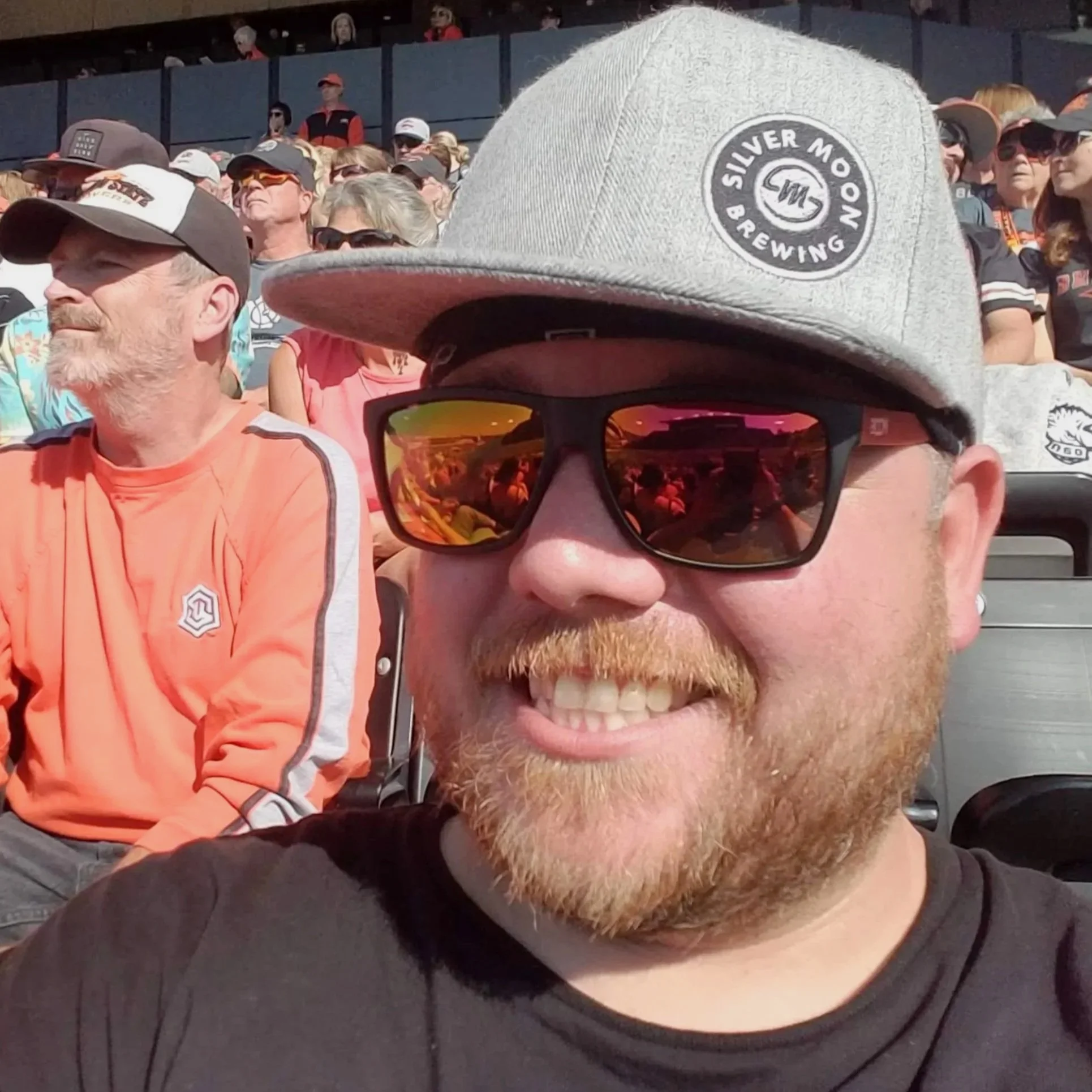 Man smiling wearing sunglasses, a gray baseball cap with a logo, and a black shirt at an outdoor event with a crowd in the background.