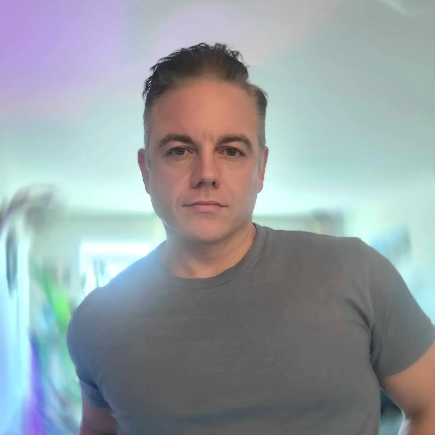 A man with short dark hair, wearing a gray T-shirt, standing in front of a blurred, colorful background.