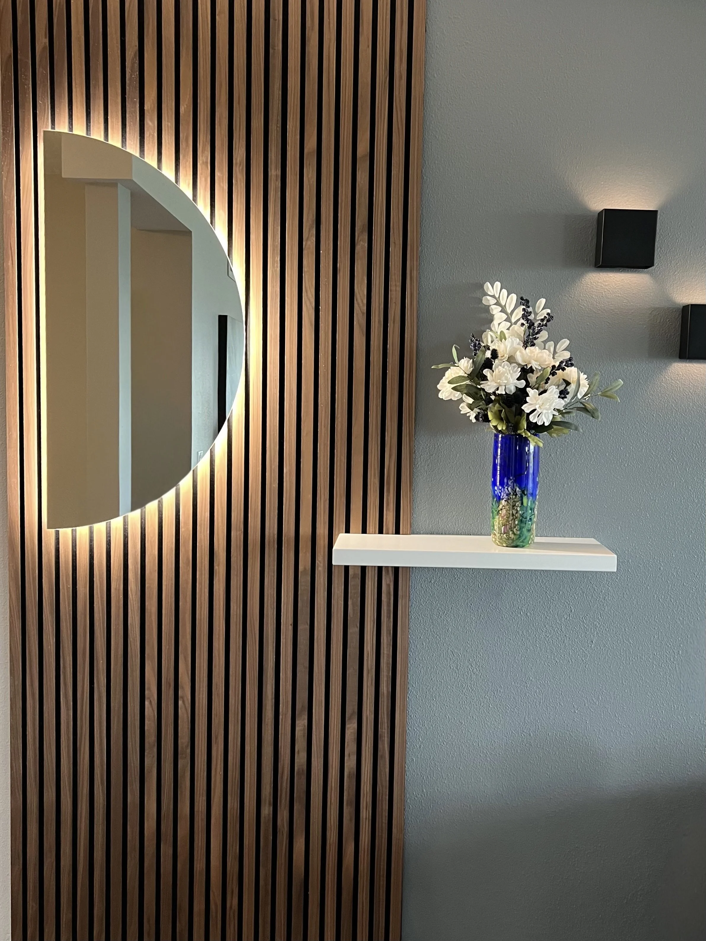 Decorative interior wall with wooden slats, a mirrored moon-shaped wall decoration with backlighting, a white shelf with a blue vase of white flowers, and two black wall-mounted lights.
