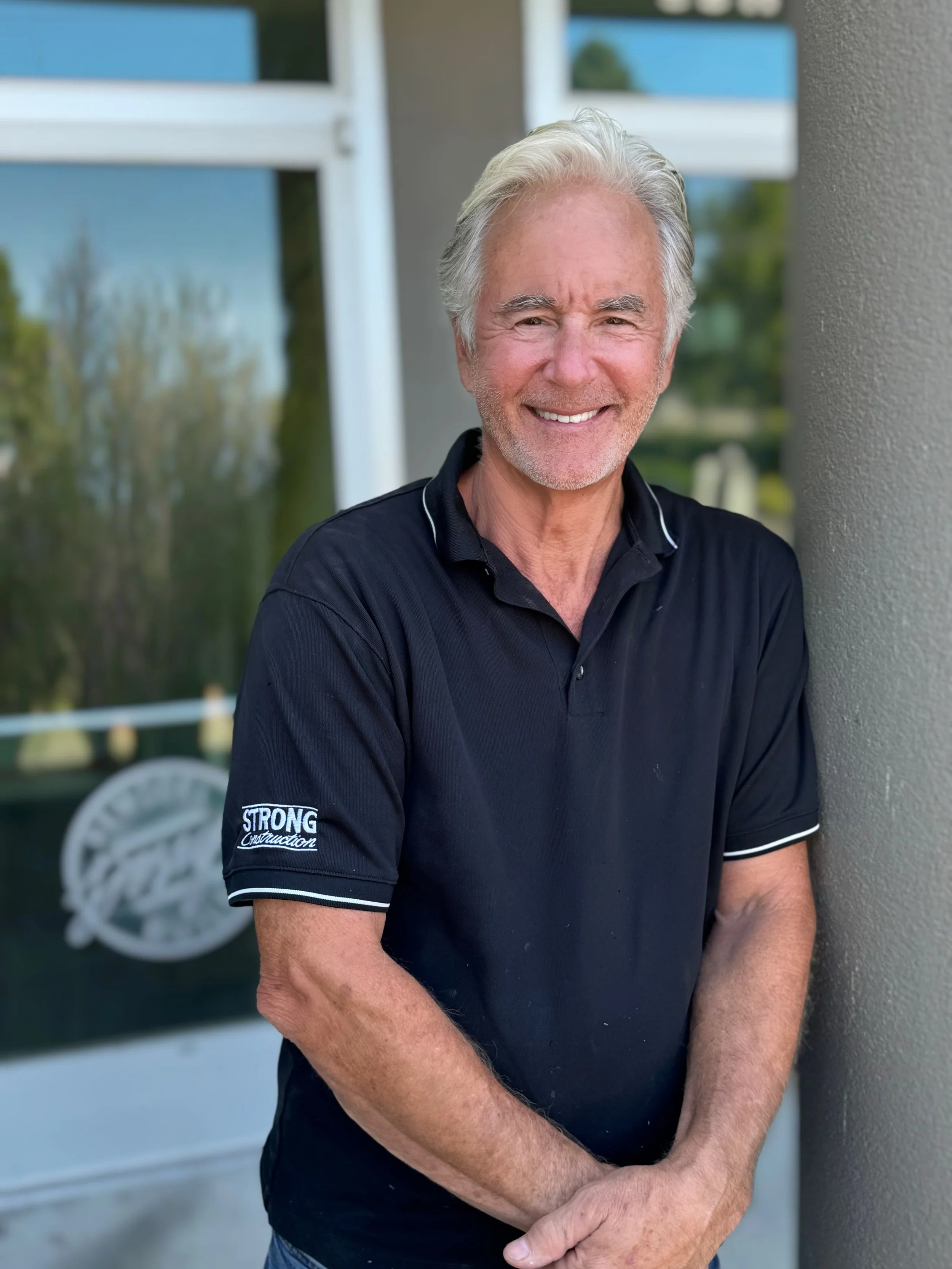A smiling elderly man with gray hair and a beard leaning against a wall outdoors, wearing a black polo shirt with white trim.