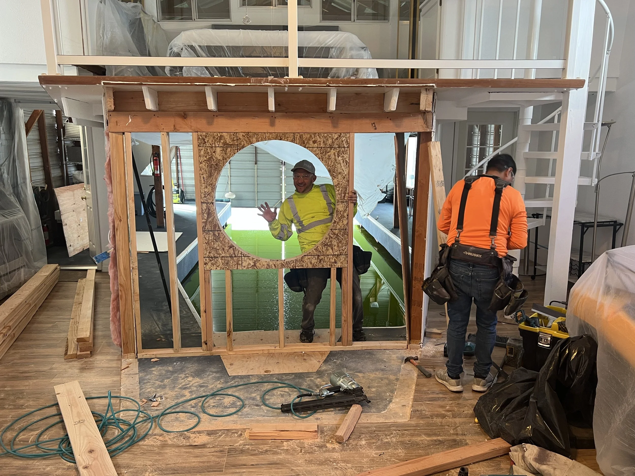 Two construction workers building a small wooden structure with a circular opening, inside a house under renovation.