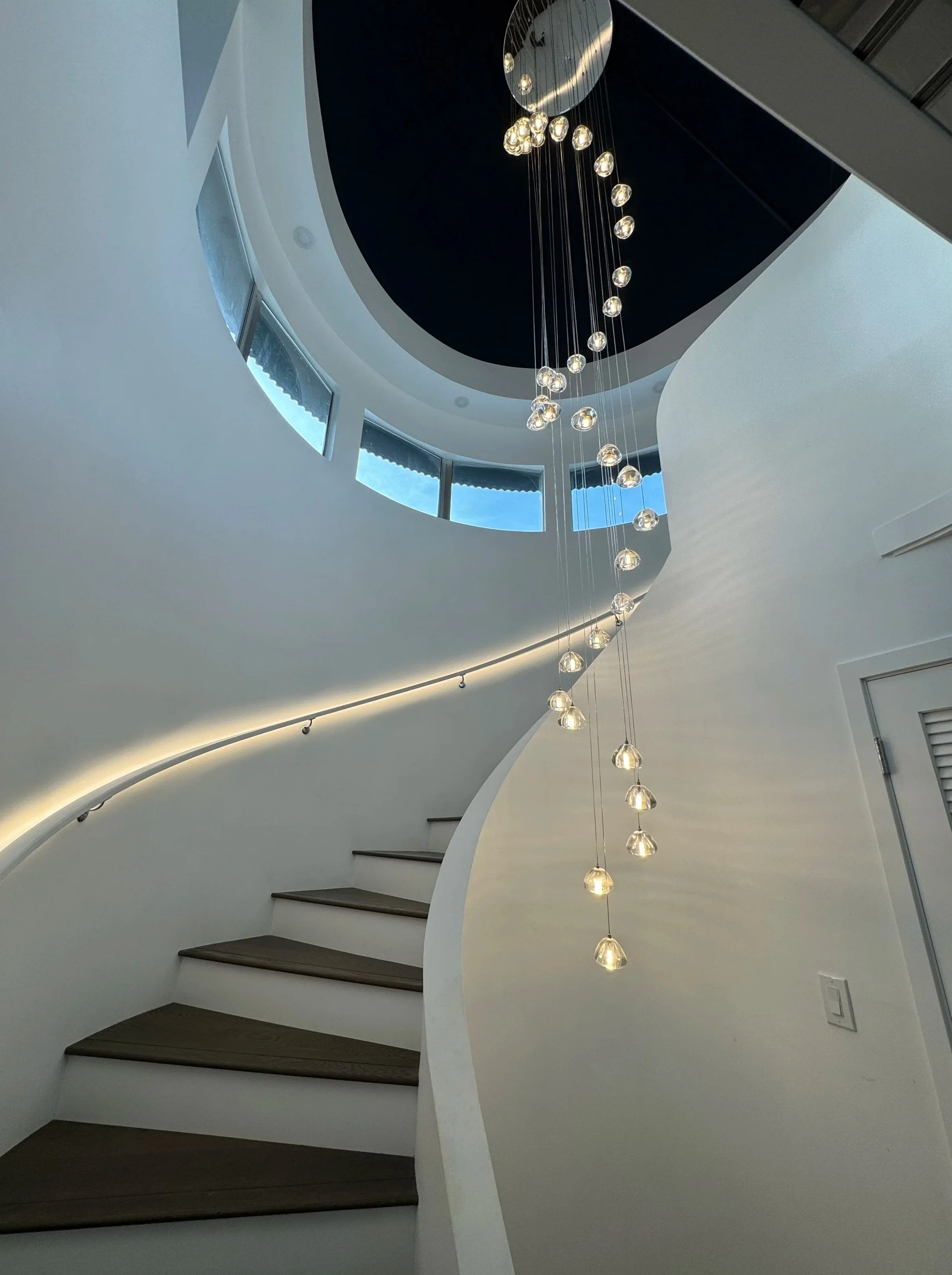 Interior view of a modern staircase with dark wooden steps and a curved white wall, a hanging chandelier with multiple glass orbs, and a circular ceiling opening with windows allowing natural light.