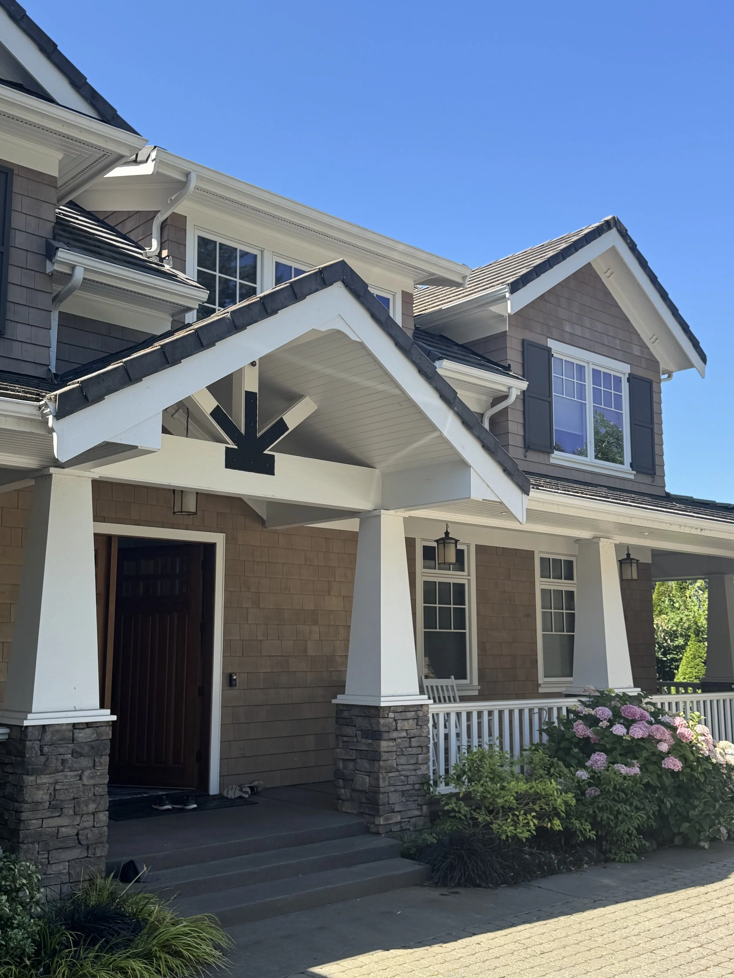 Front view of a two-story house with a porch, flower bushes, and a clear blue sky.