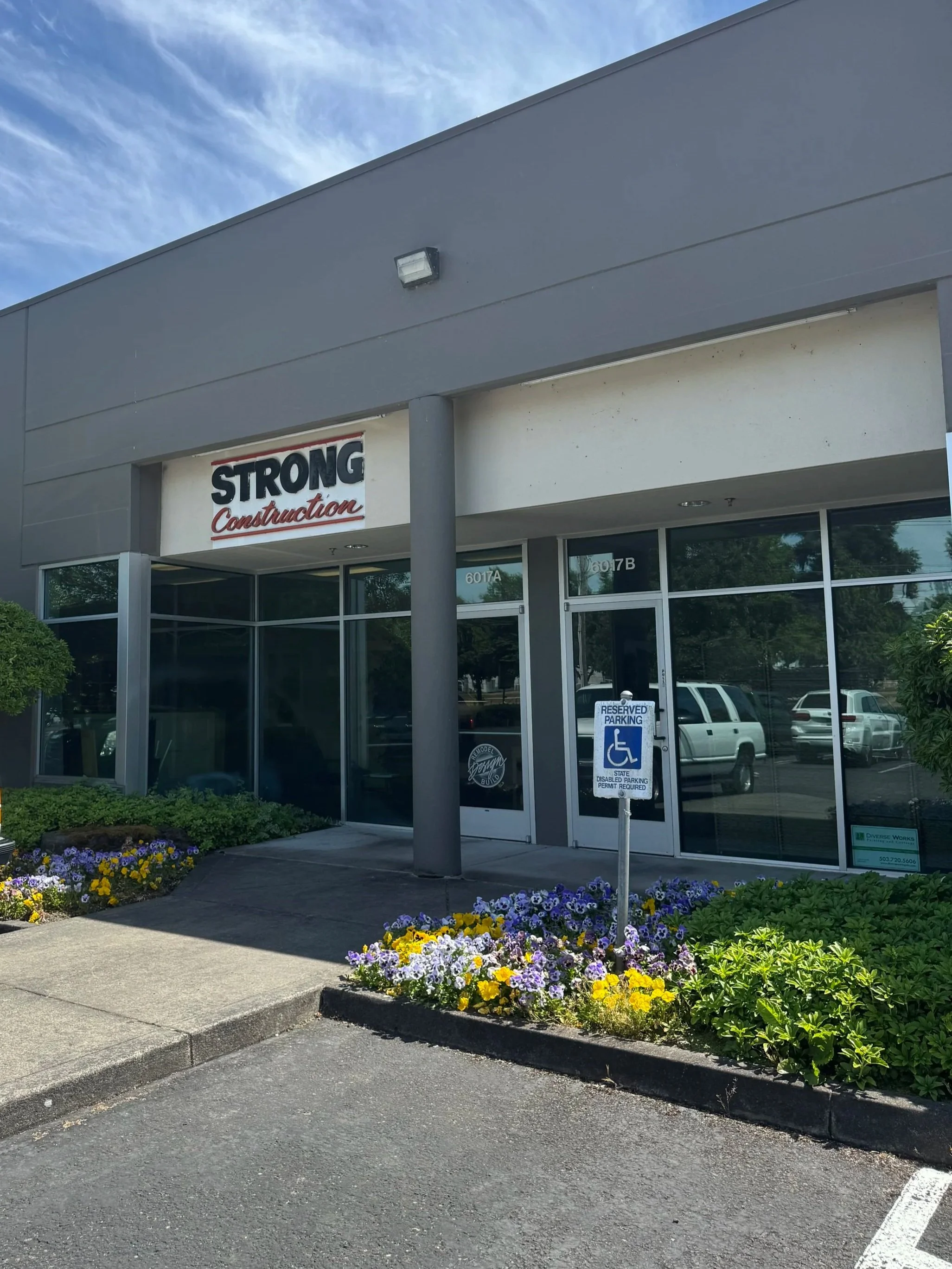 Front of a commercial building with a sign that reads 'Strong Construction.' There is a reserved parking spot for people with disabilities and a small, landscaped area with colorful flowers in front.