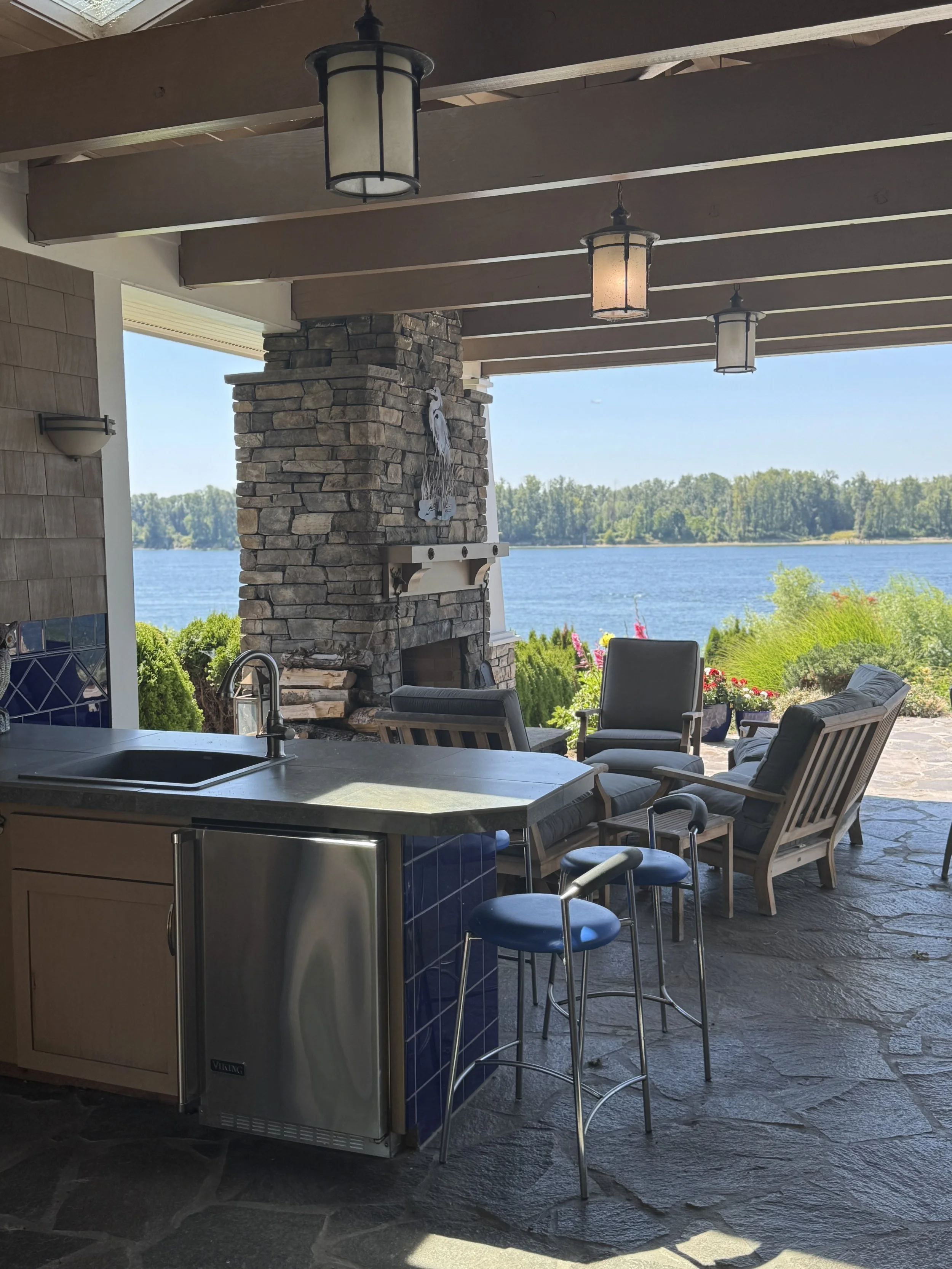 Outdoor patio with stone fireplace, wooden ceiling beams with hanging lanterns, and a view of a lake with trees in the background. There is a kitchen counter with bar stools and a seating area with outdoor chairs and a sofa.