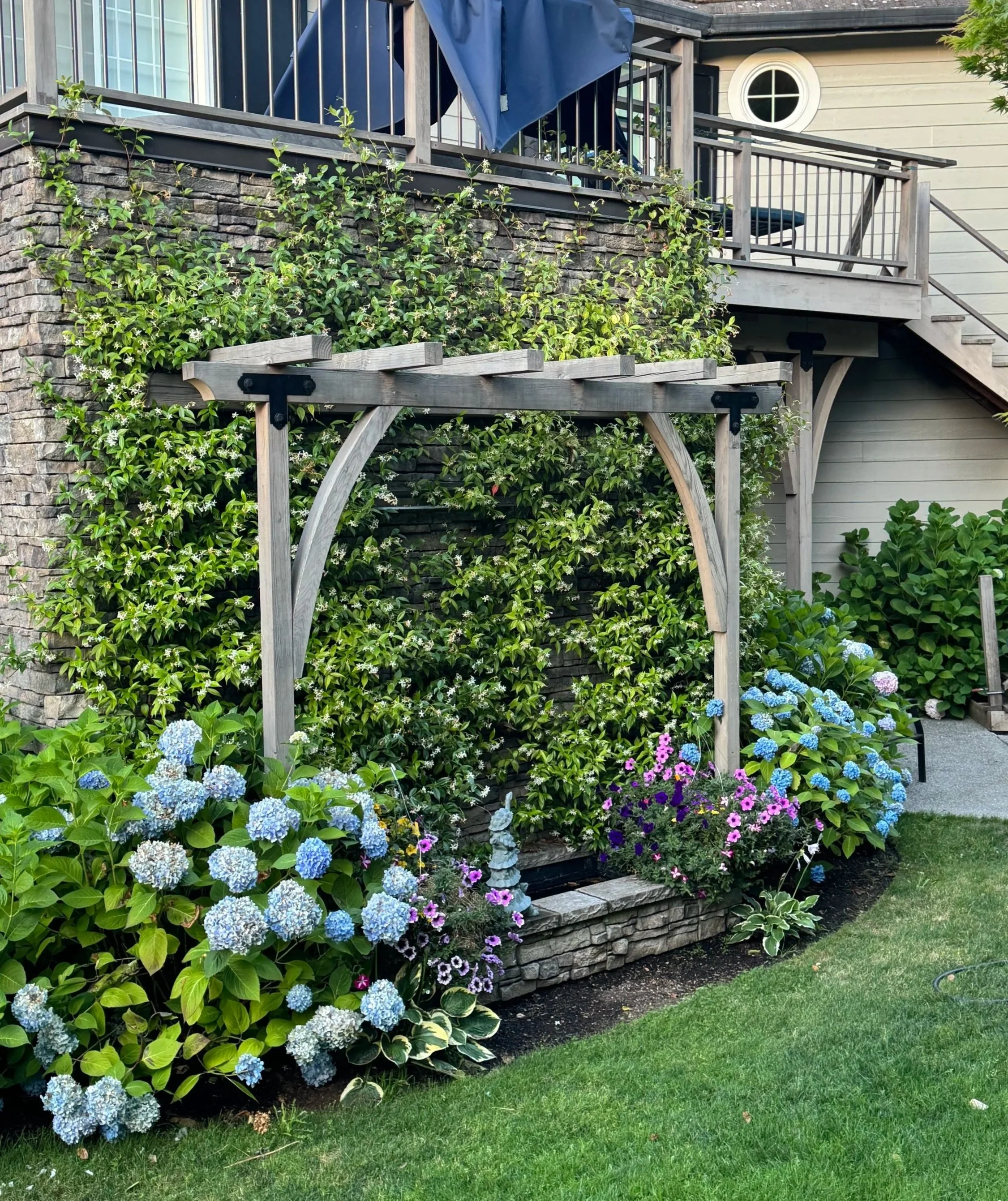A garden fenced with a brick wall and lush green plants, with colorful hydrangeas and small flowers in front, and a wooden arbor frame.
