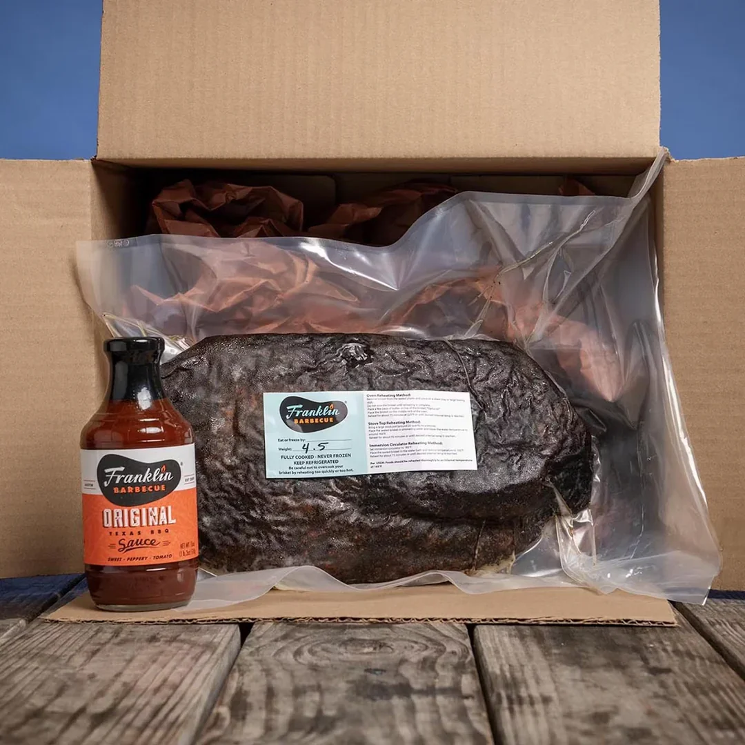 A large frozen smoked brisket in vacuum packaging inside a cardboard box, with a bottle of Franklin Barbecue original barbecue sauce placed next to it on a wooden surface.