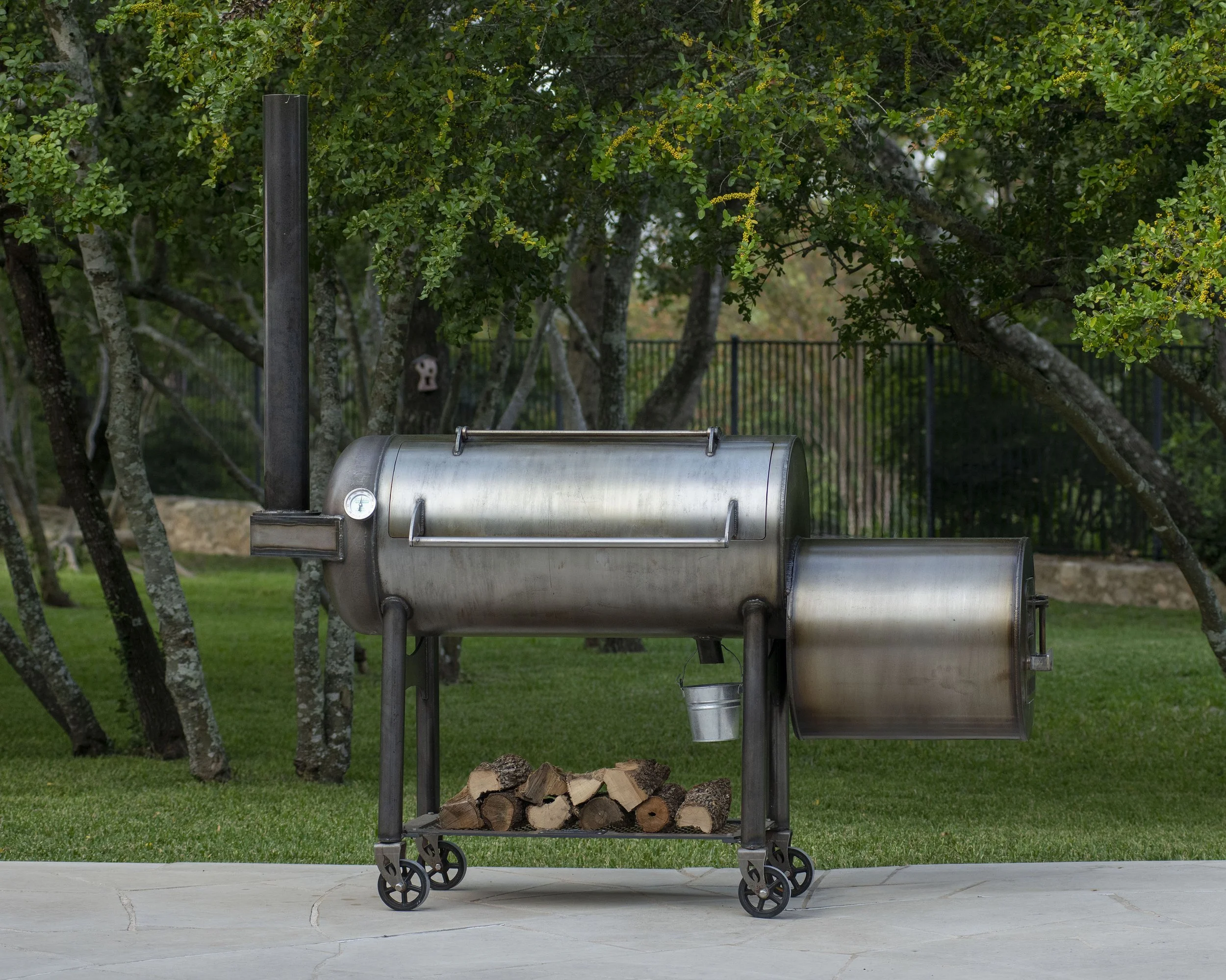 A backyard scene with a Franklin Barbecue Pit Smoker on wheels, filled with chopped firewood, standing on a concrete patio surrounded by grass and trees with green leaves and yellow flowers.