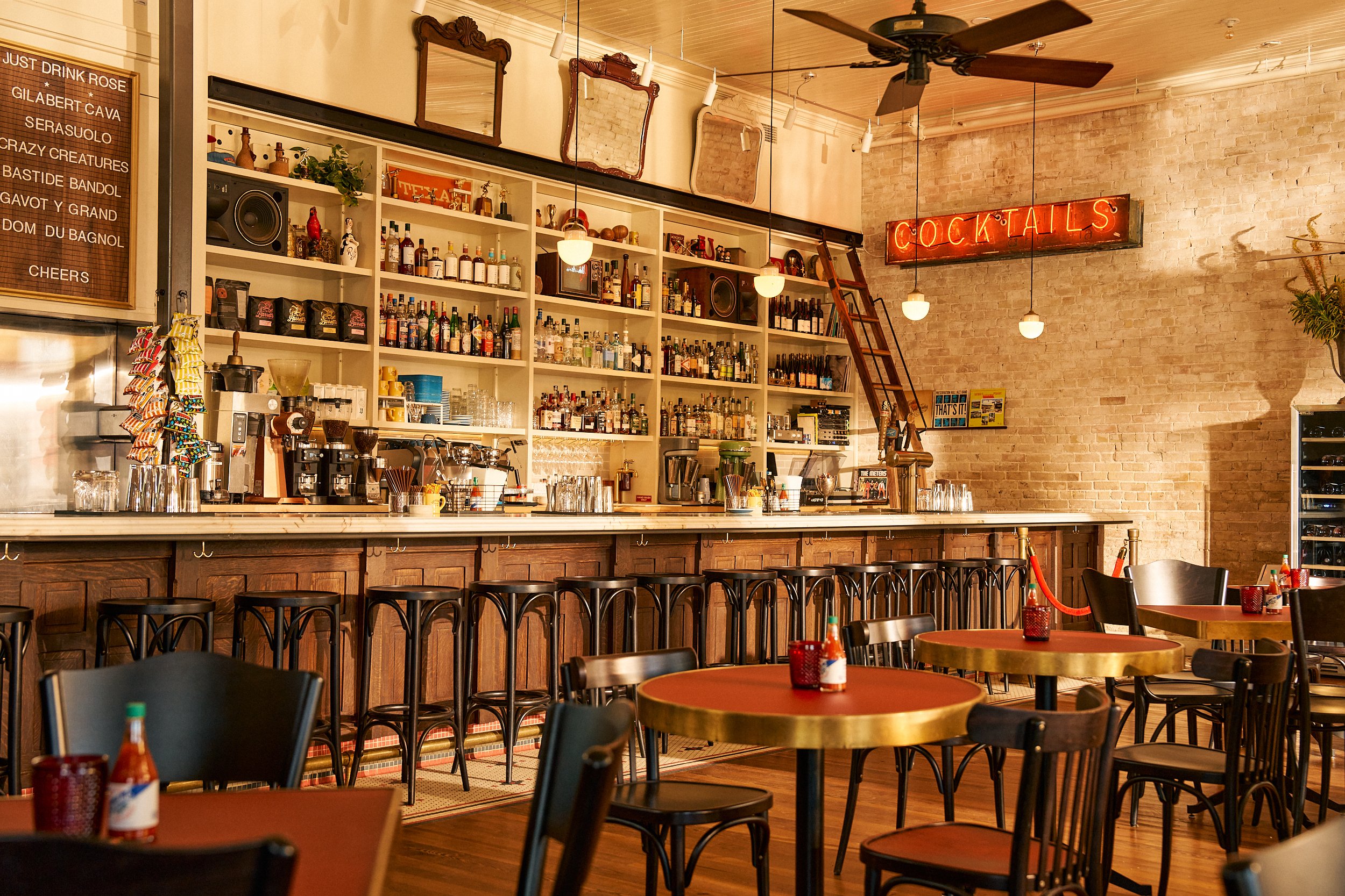 Interior of a cozy bar or cafe with a long counter, bar stools, round wooden tables with chairs, a brick wall, ceiling fan, and a neon "Cocktails" sign.