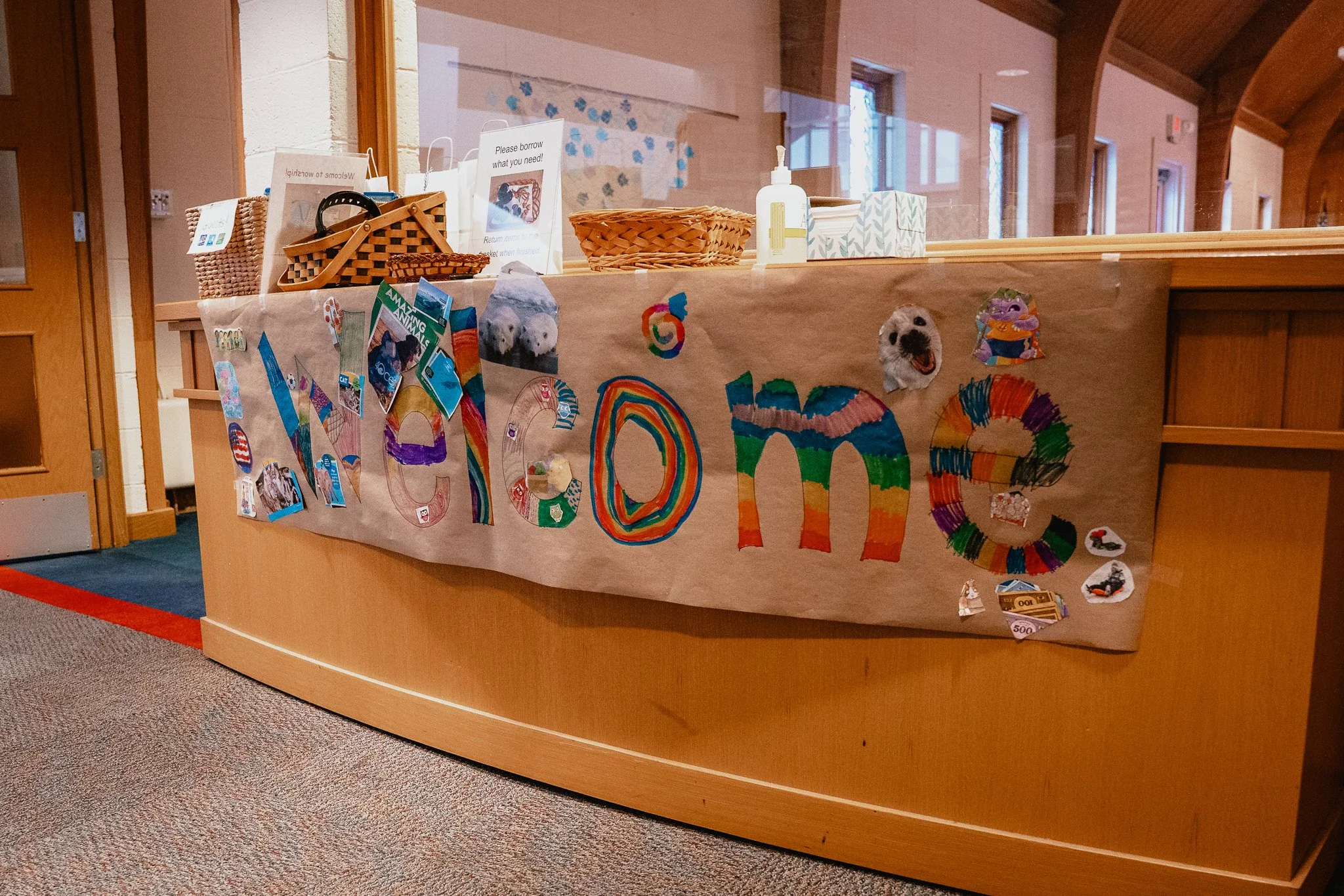 Library or community center desk with a colorful banner that spells 'Welcome' made from decorated letters. The counter holds baskets, signs, and hand sanitizer, with windows and wooden architecture in the background.
