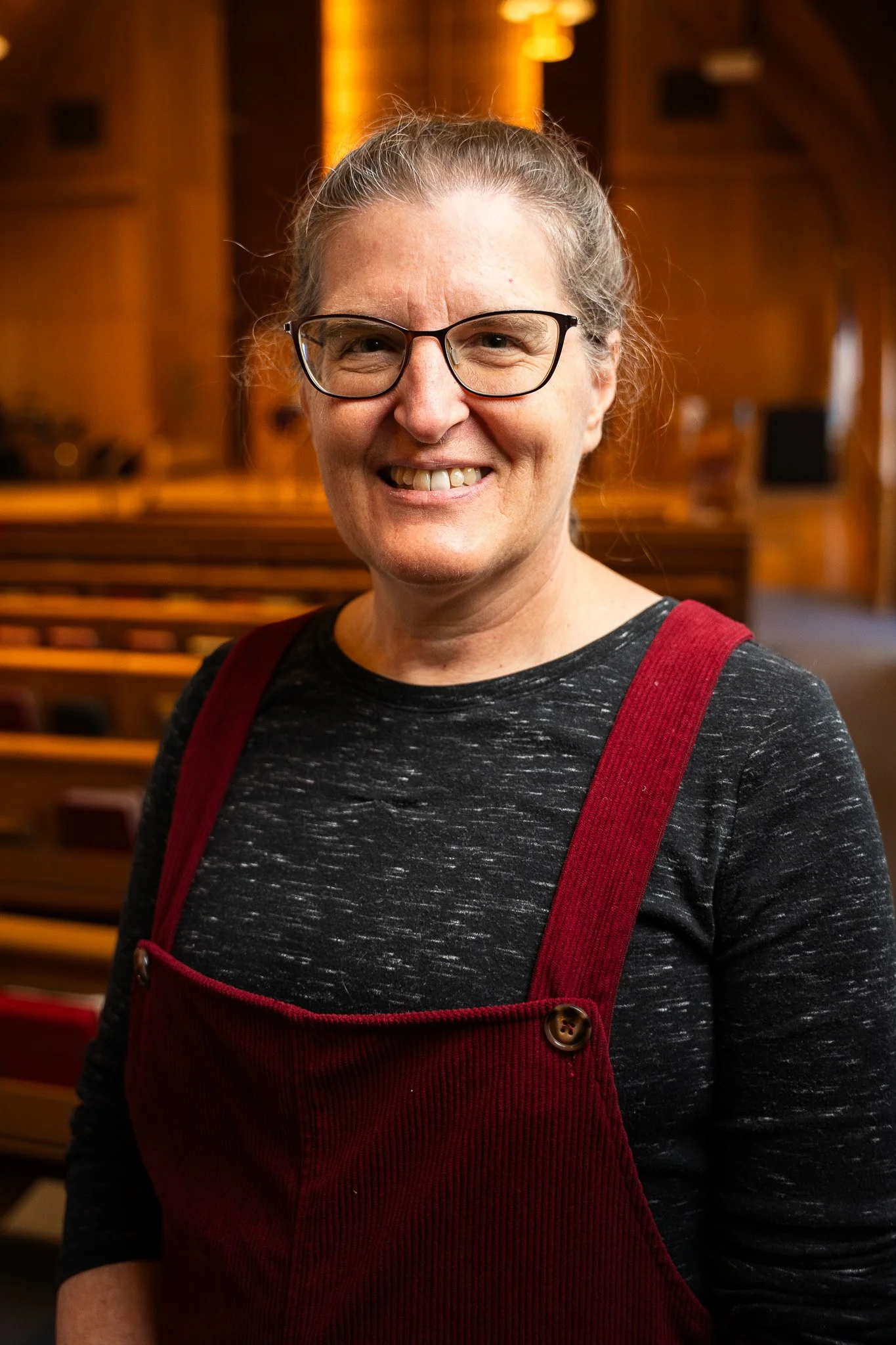 A smiling woman with glasses, gray hair, wearing a black mottled shirt and red overalls standing inside a warmly-lit wooden church or hall.
