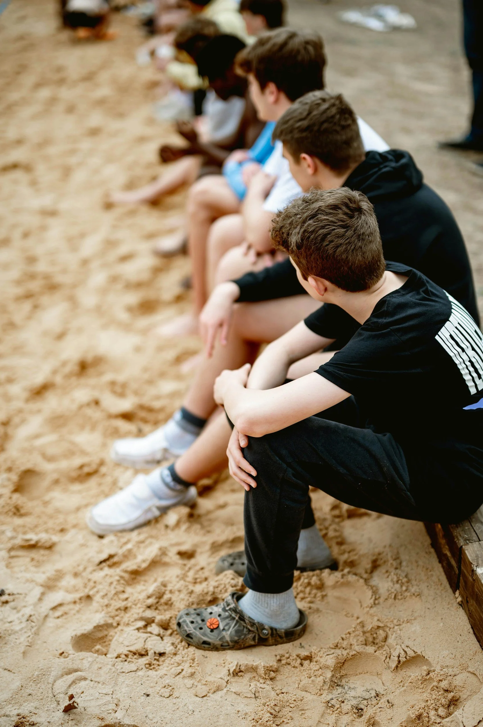 A group of young boys sitting on a bench with their feet on sandy ground, with some sporting sneakers and Crocs, looking in the same direction, possibly watching or listening to something.