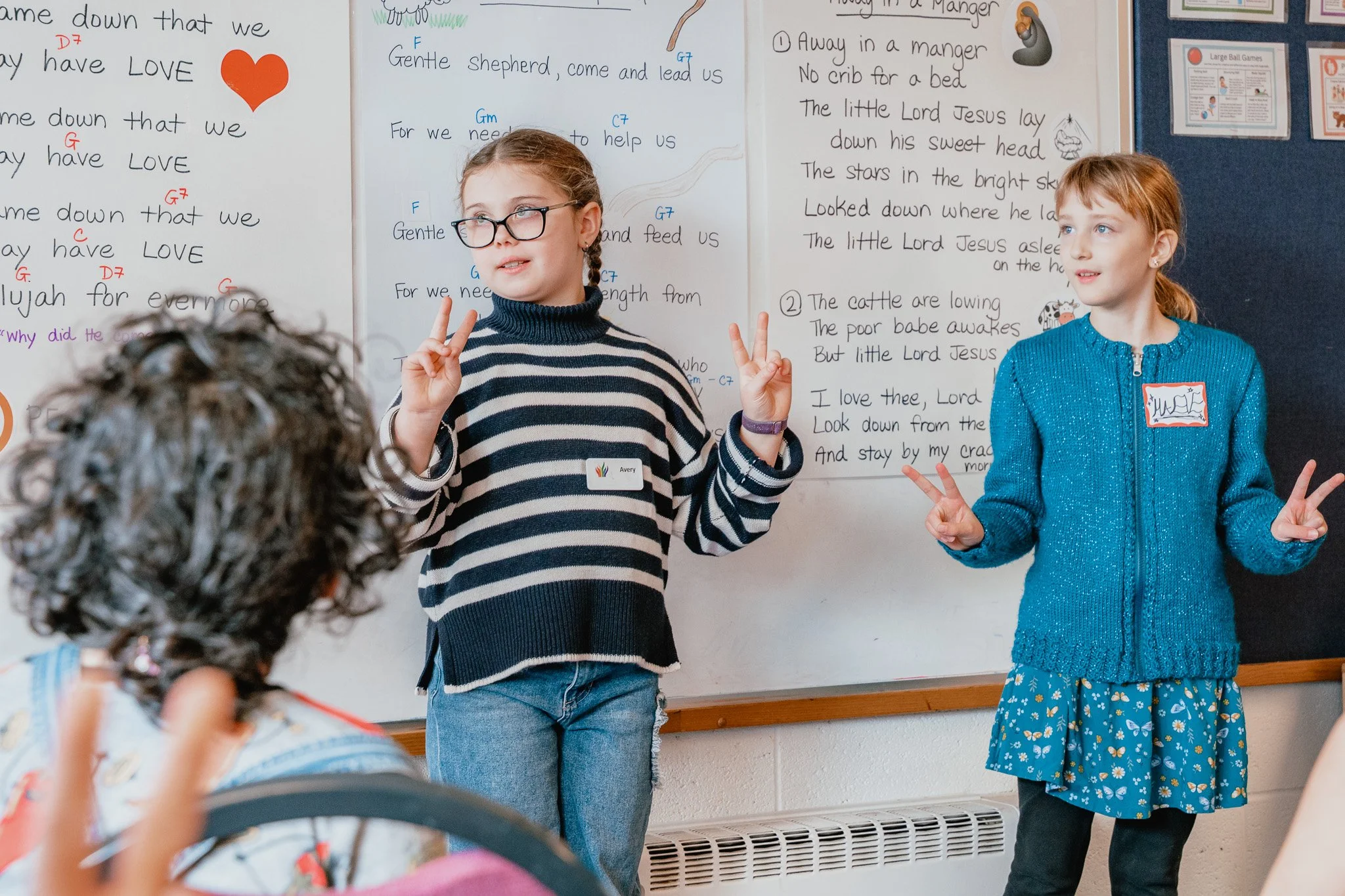 Two young girls standing in front of a whiteboard, making peace signs with their hands during a classroom presentation. The girl on the left has glasses and braided hair, wearing a striped sweater, while the girl on the right has red hair and a blue jacket. The whiteboard behind them is filled with handwritten lyrics, musical notes, and colored markings.