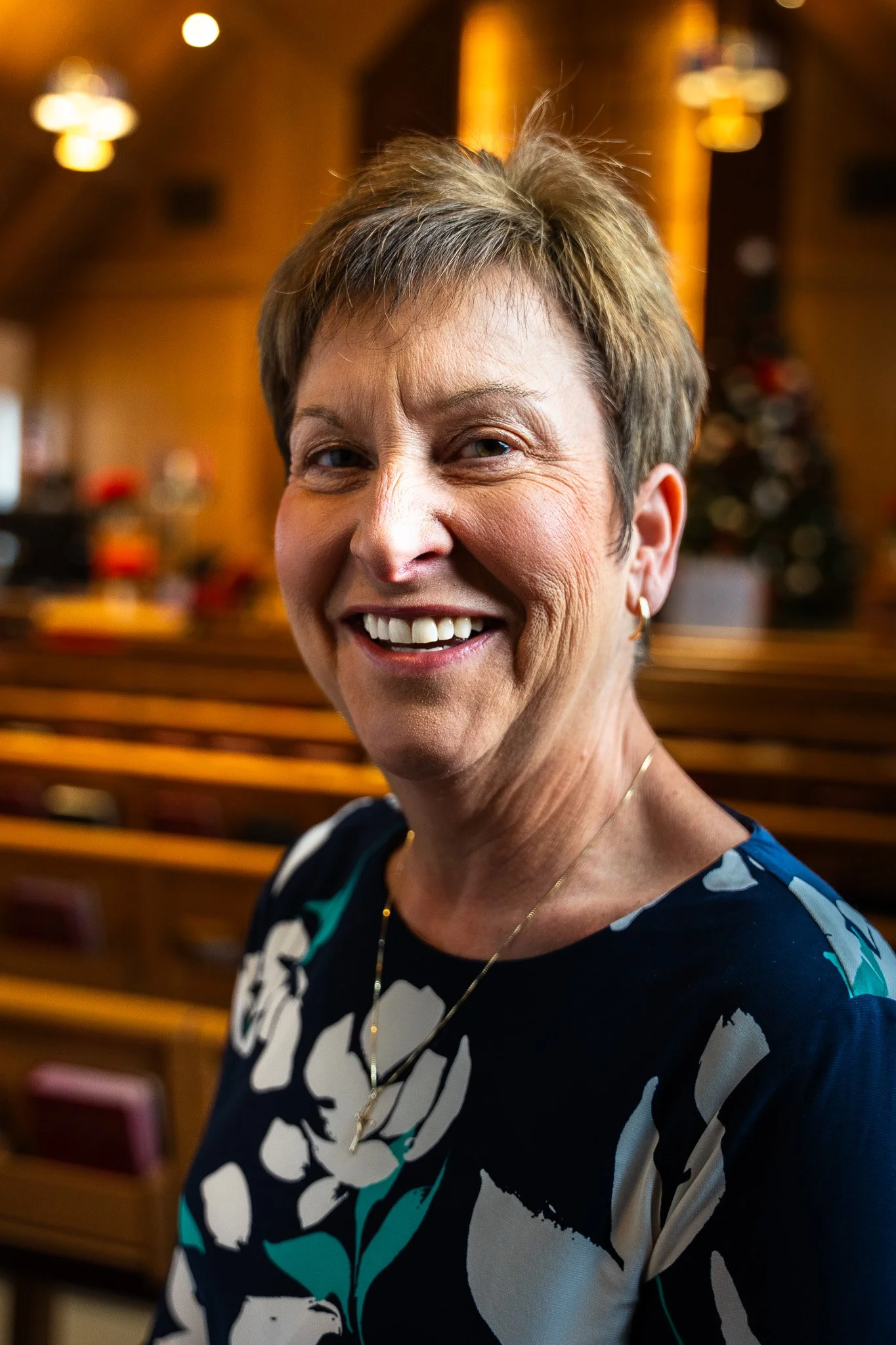 Portrait of a middle-aged woman smiling, wearing a black top and a gold necklace, inside a wooden-paneled room with warm lighting.