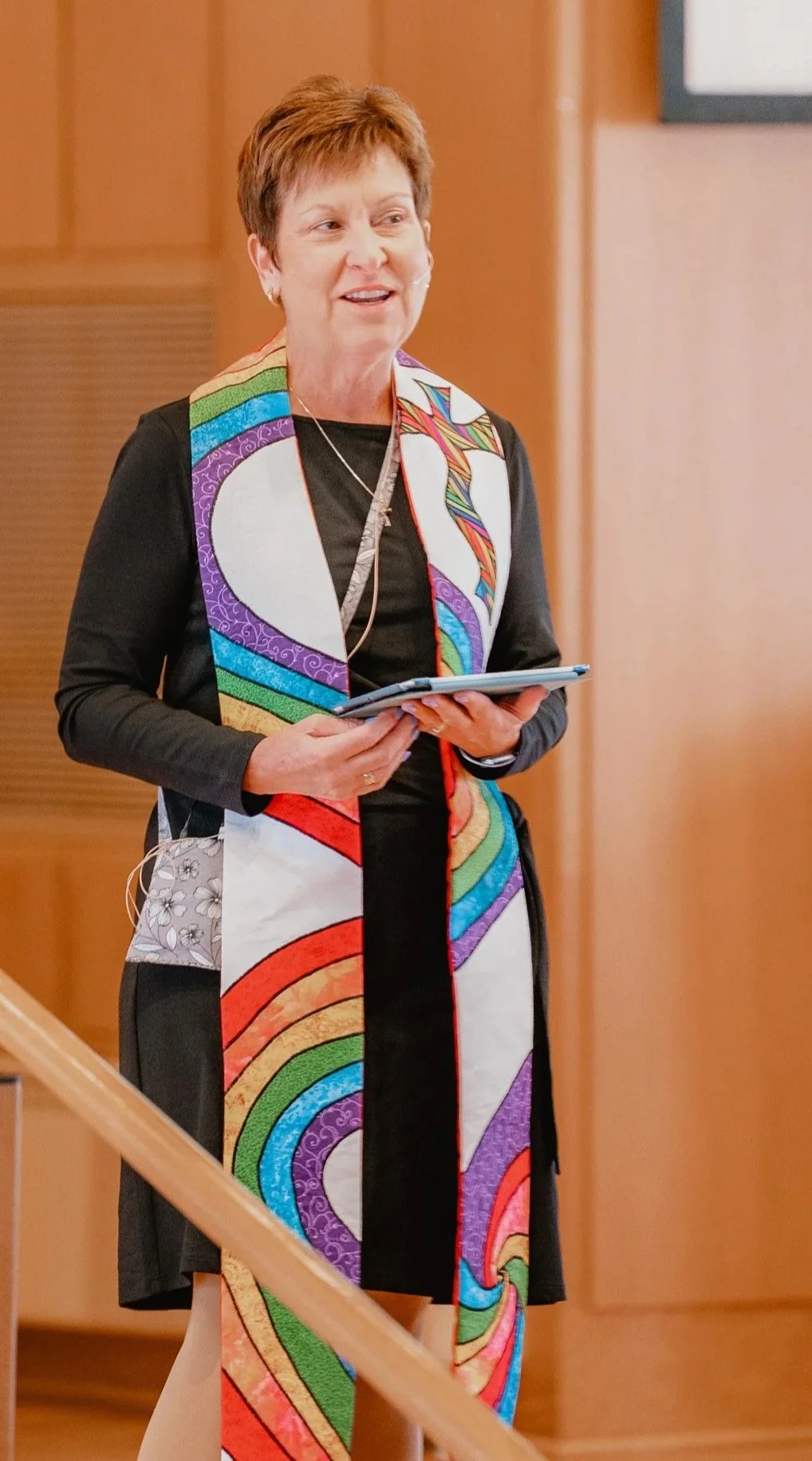 A woman with short reddish hair wearing a colorful, patterned
vest holds a tablet or digital device, and appears to be speaking during a presentation or discussion in a professional setting.