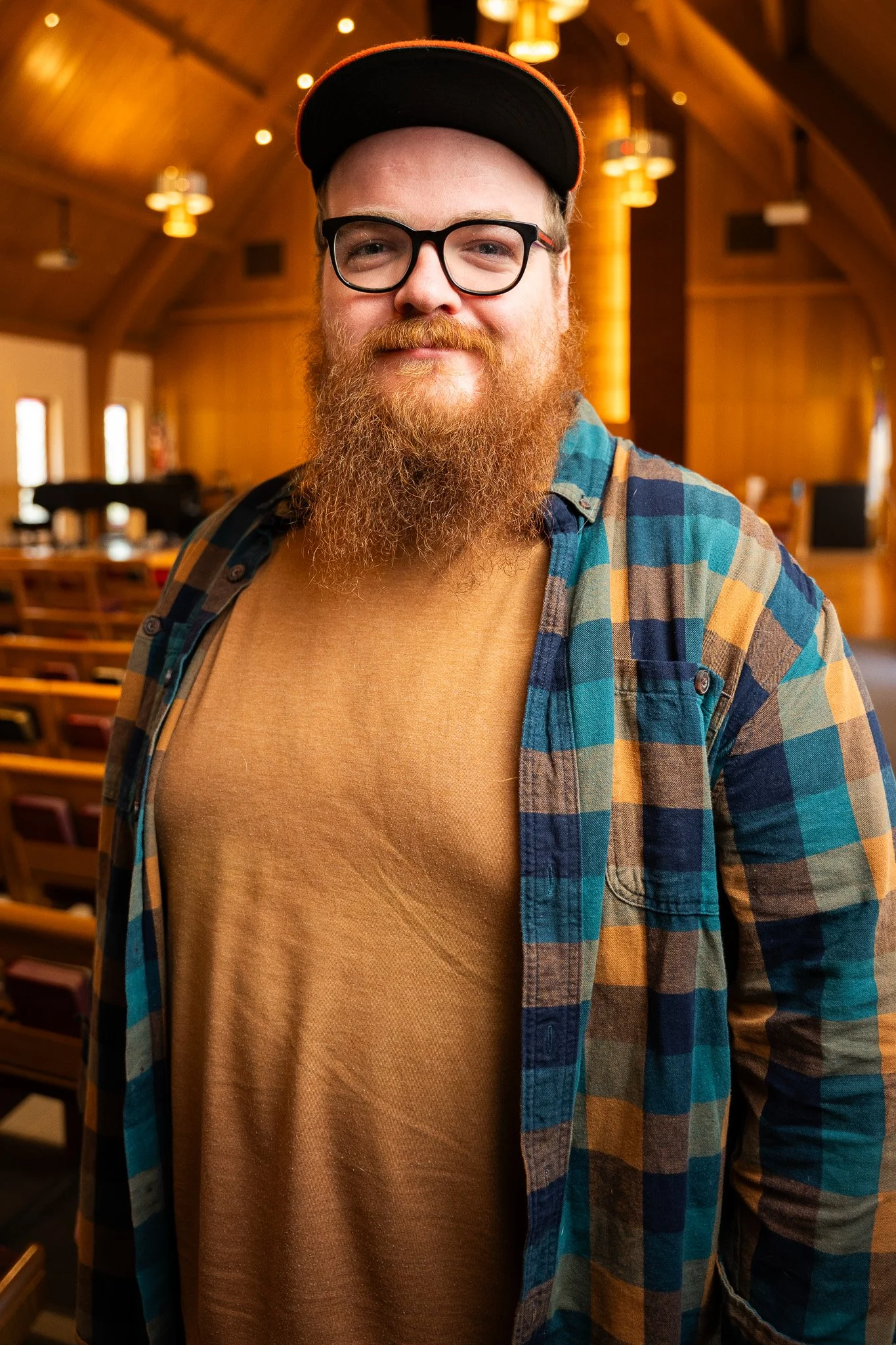 A man with glasses, a red beard, and a baseball cap smiling inside a wooden hall with rows of chairs.
