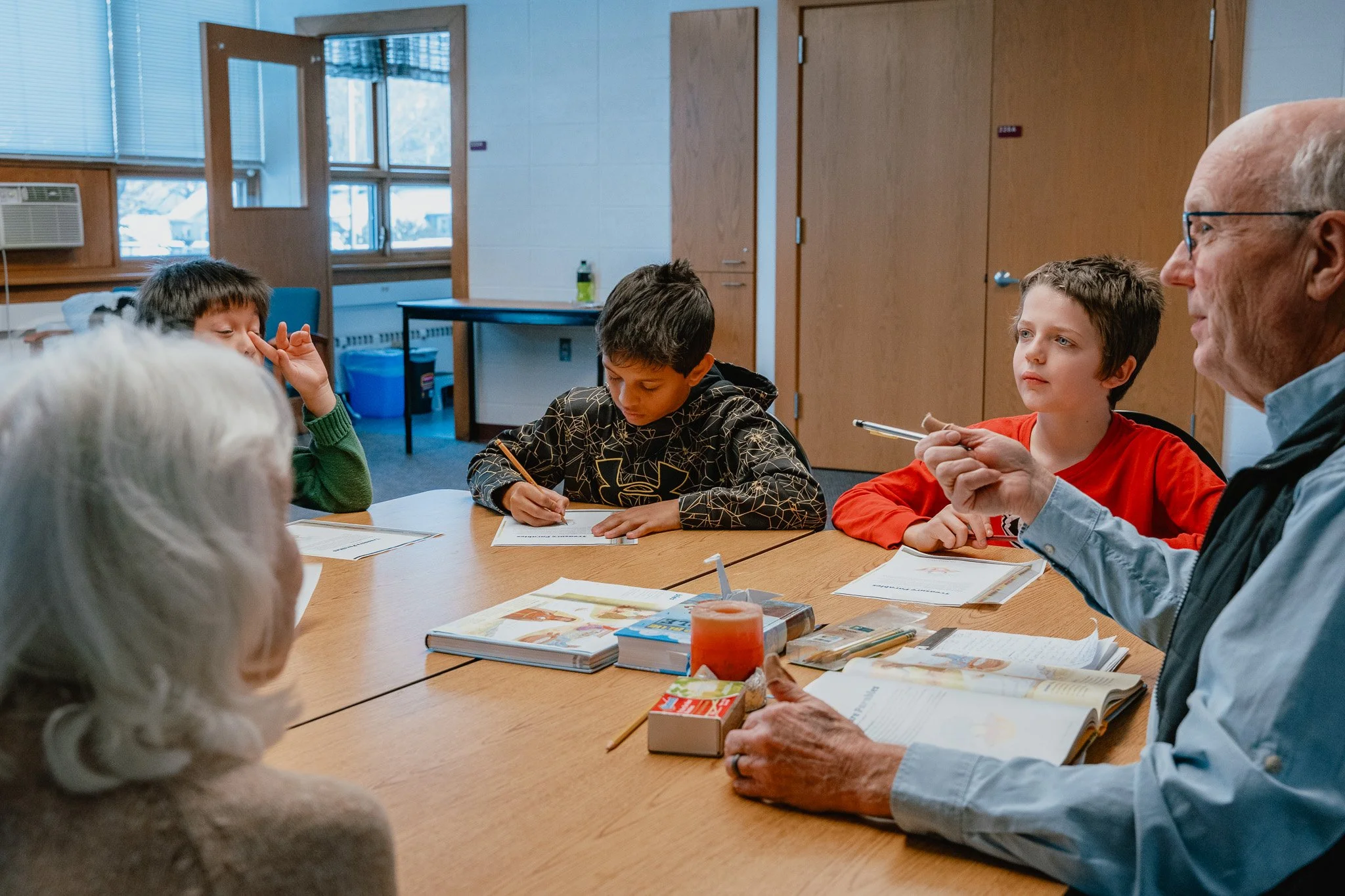 A group of children and an adult sit around a wooden table in a classroom or library, engaged in a discussion or lesson. The table has books, papers, and a candle on it. The room has large windows, wooden doors, and a whiteboard in the background.