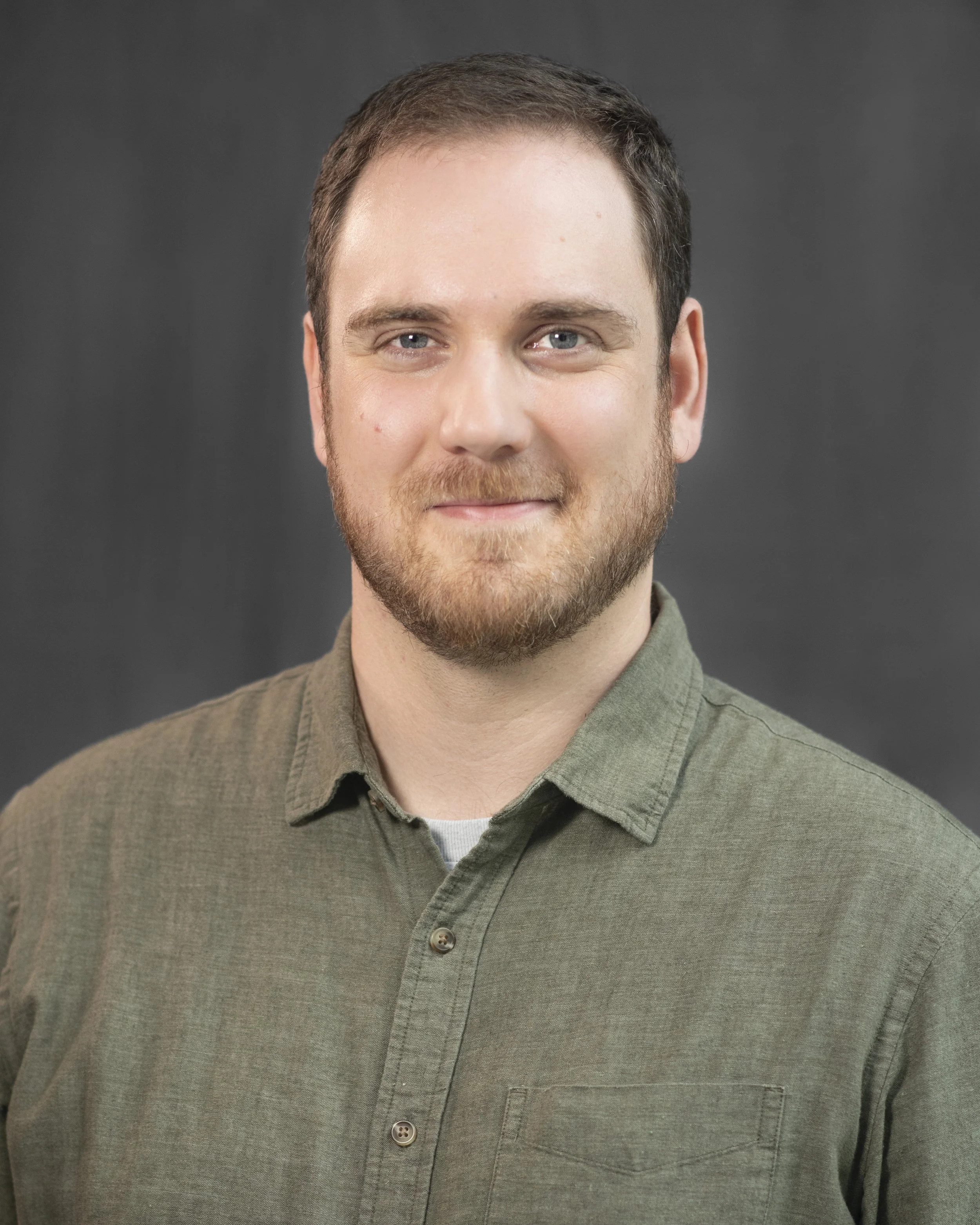 A smiling man with a beard and short hair standing inside a wooden church or hall, wearing a green sweater with a zip collar.