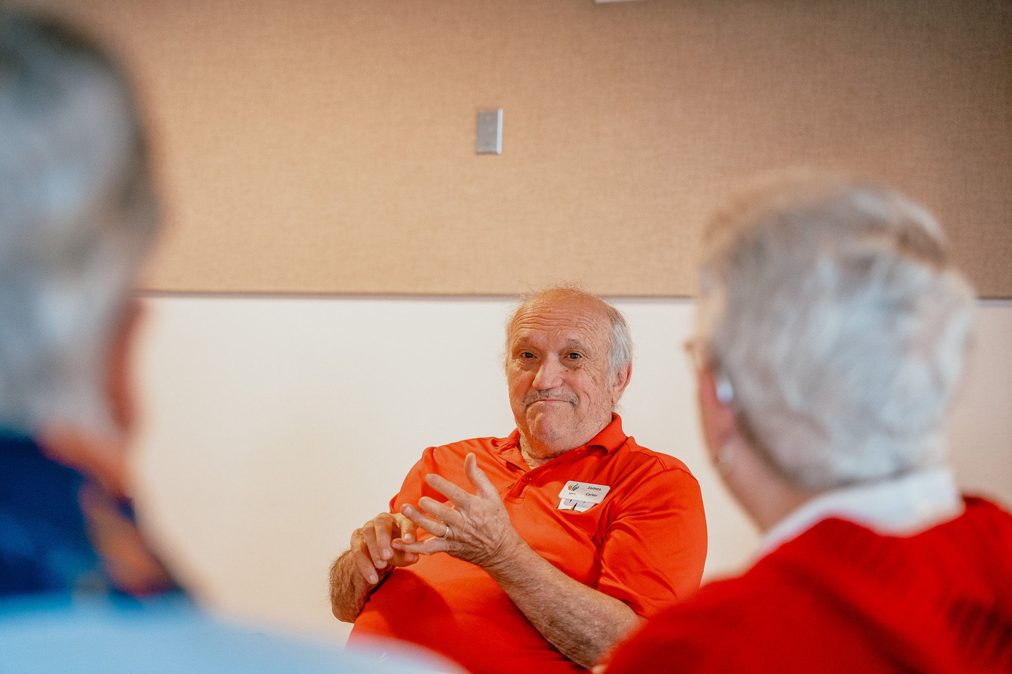 An older man in a red shirt speaking to two people in a room with beige walls.