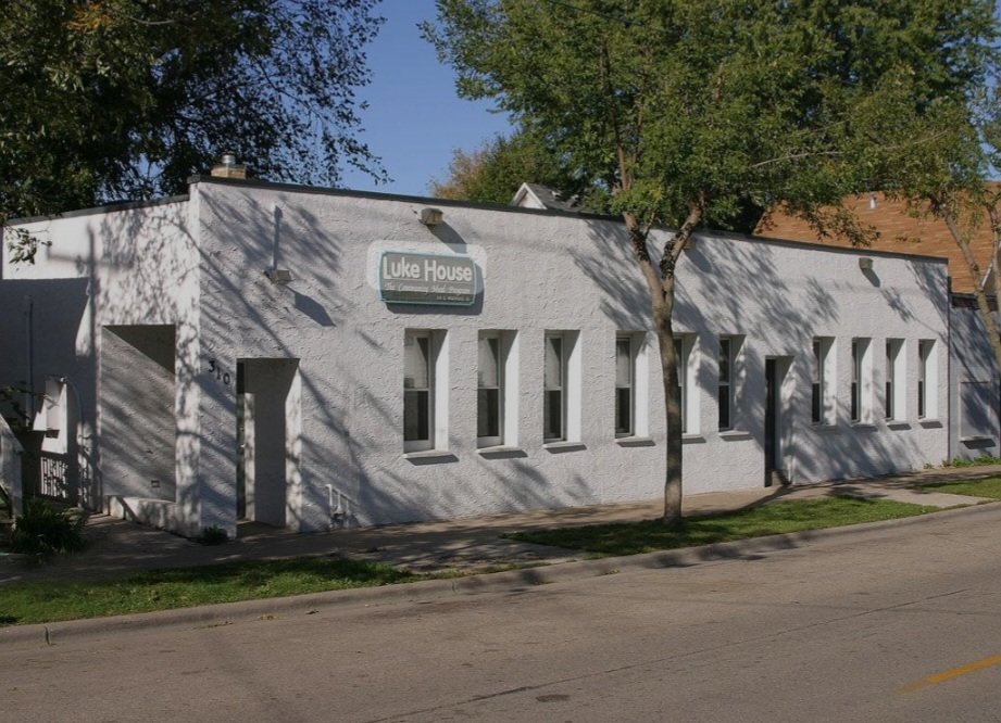 A white building with a sign that reads "Luke House" on the exterior wall, with a sidewalk in front and a tree casting shadows on the building, located on a street in a suburban neighborhood.