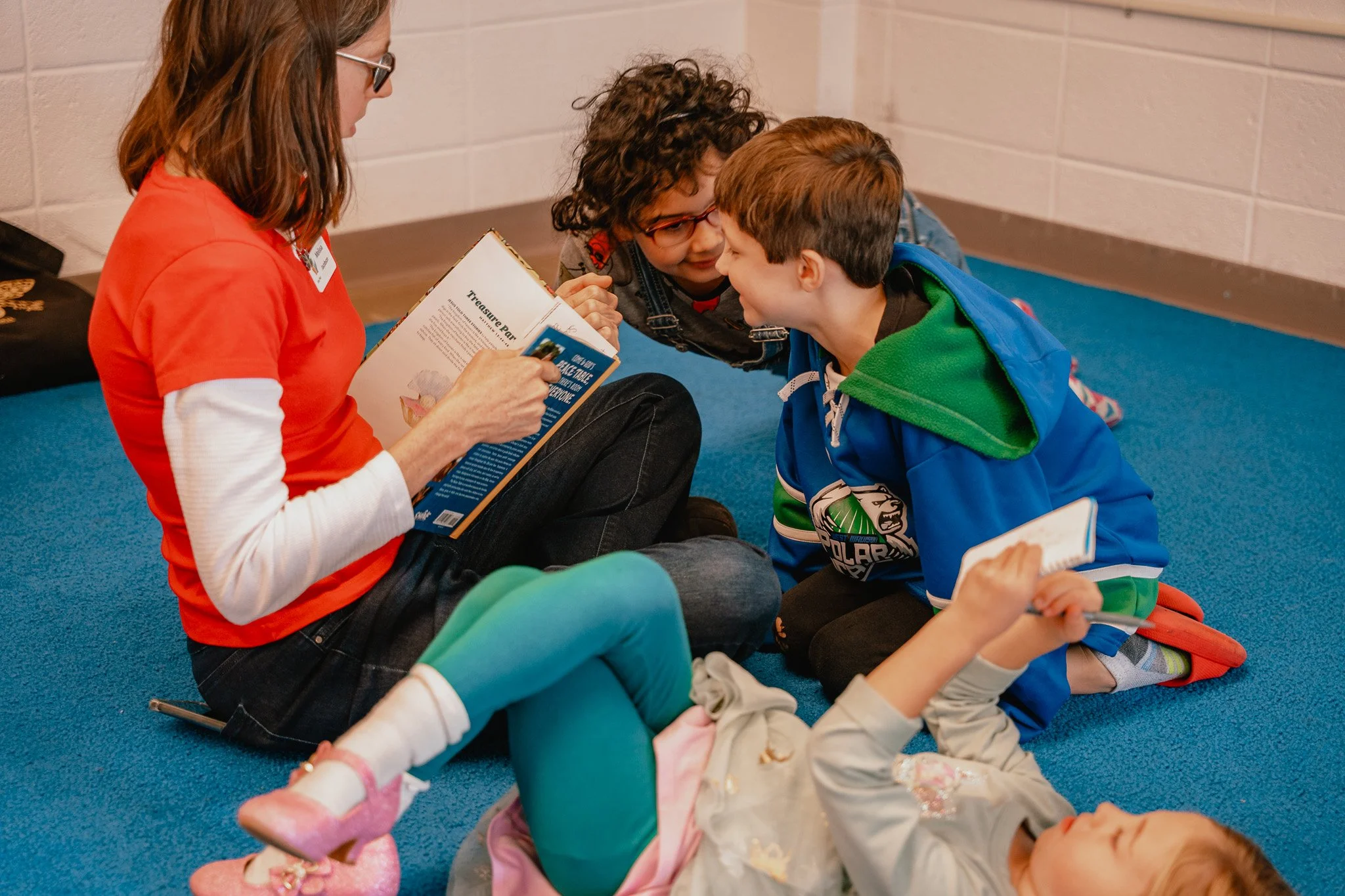 A woman reading a book to three children on a blue carpeted floor in a classroom or library setting.