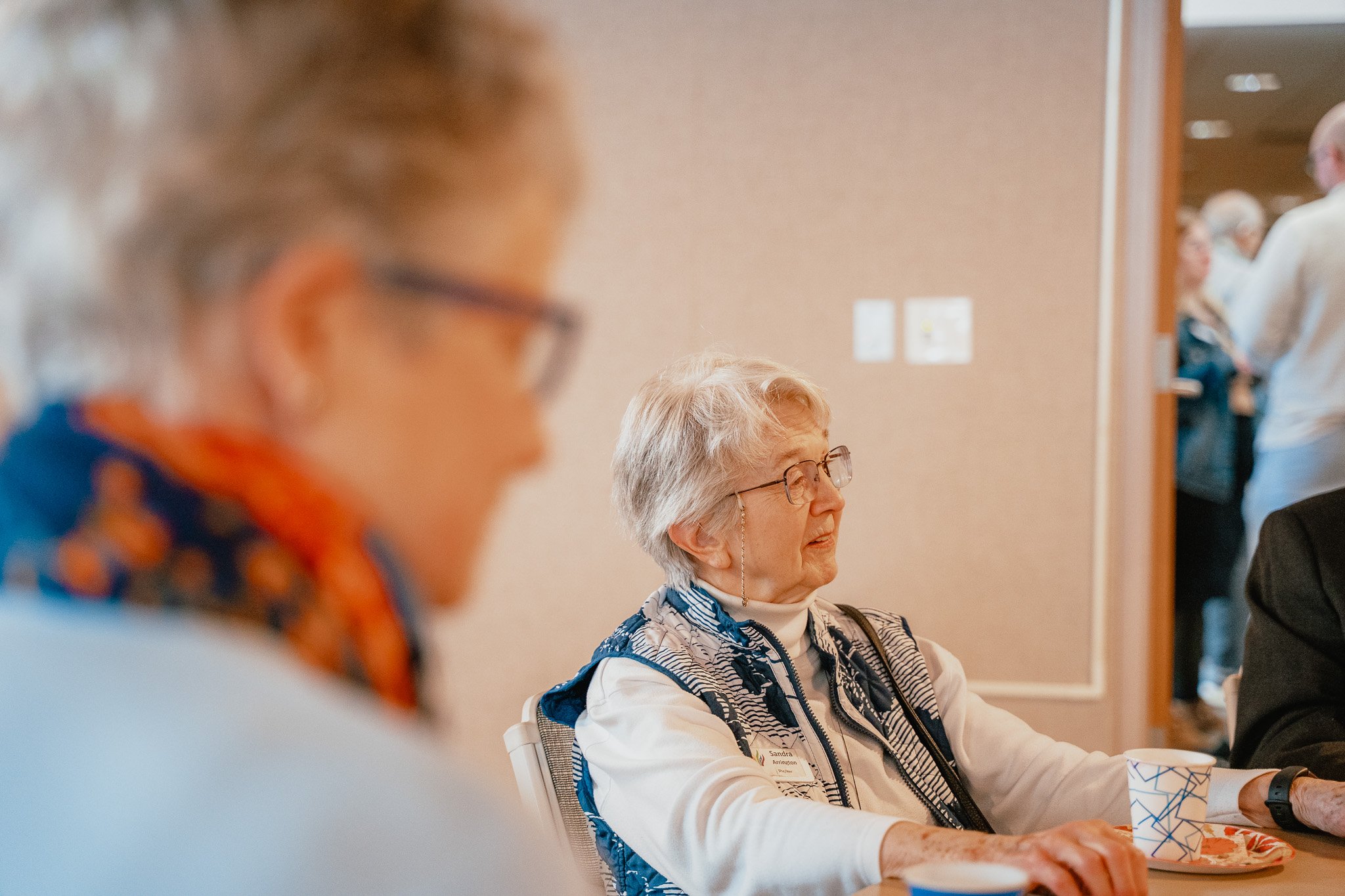 An elderly woman with glasses and light-colored hair sitting at a table with a paper cup and a plate, in an indoor setting with other people in the background.