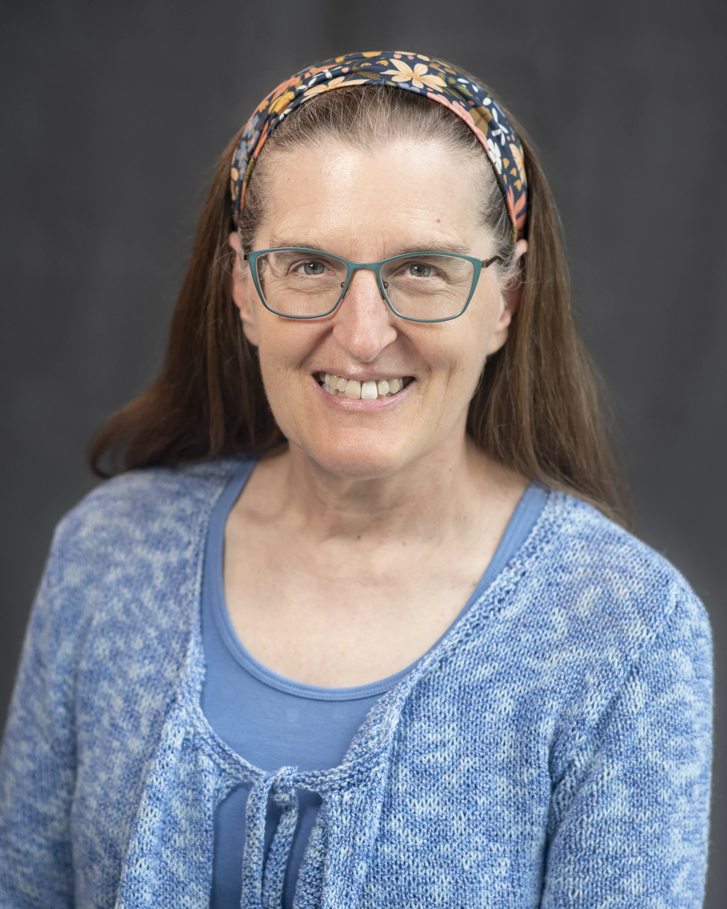A smiling woman with glasses, gray hair, wearing a black mottled shirt and red overalls standing inside a warmly-lit wooden church or hall.