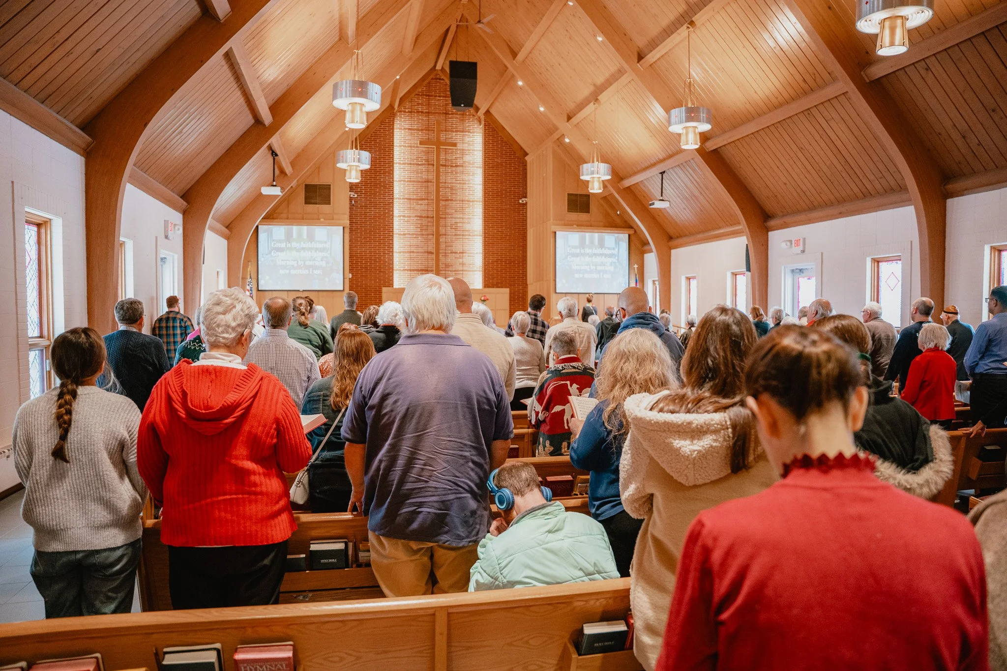 People gathered inside a church during service, standing at pews facing the front where a cross and screens are visible.