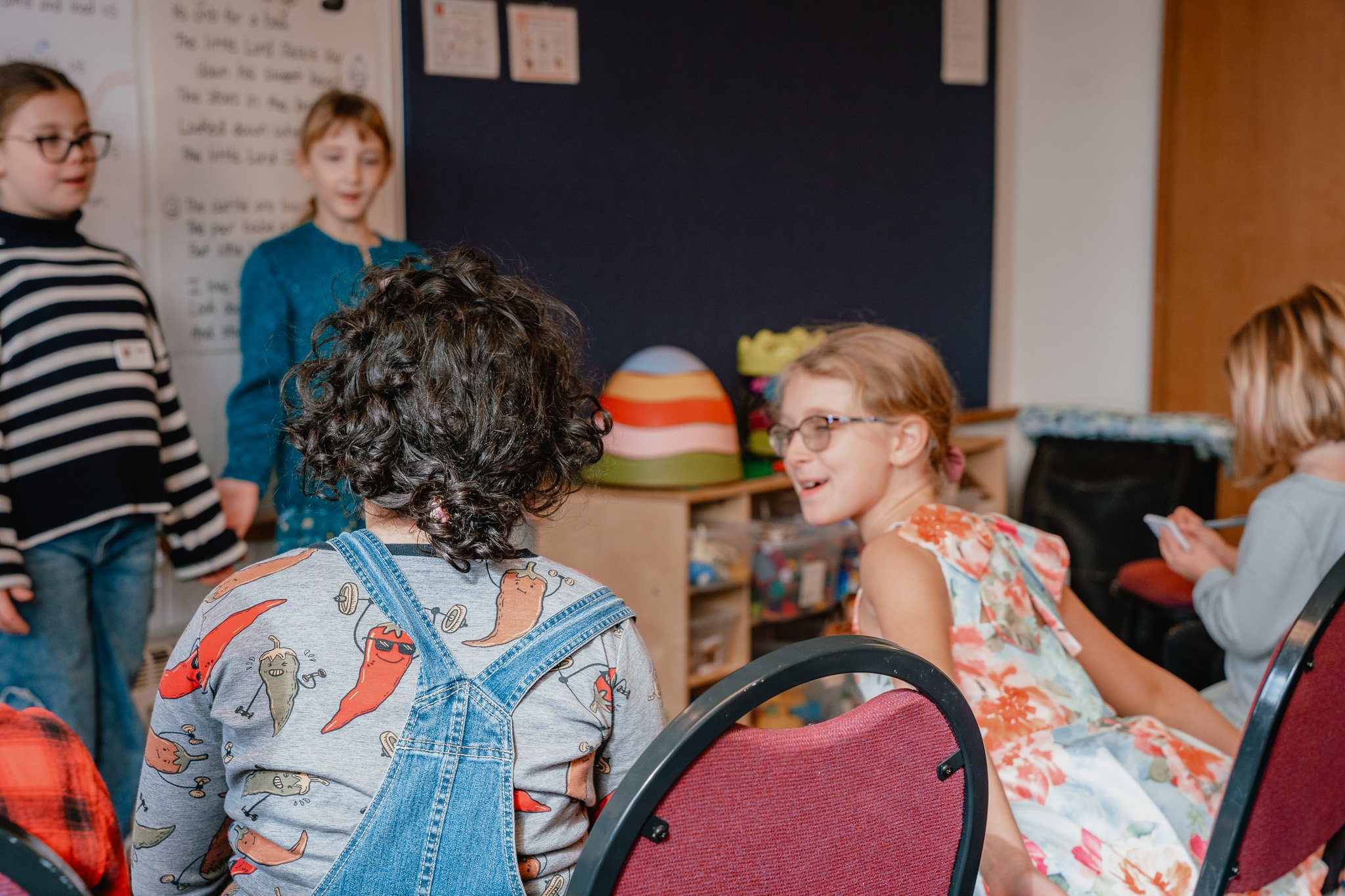 Children in a classroom, some standing and some sitting, engaged in conversation and activities.