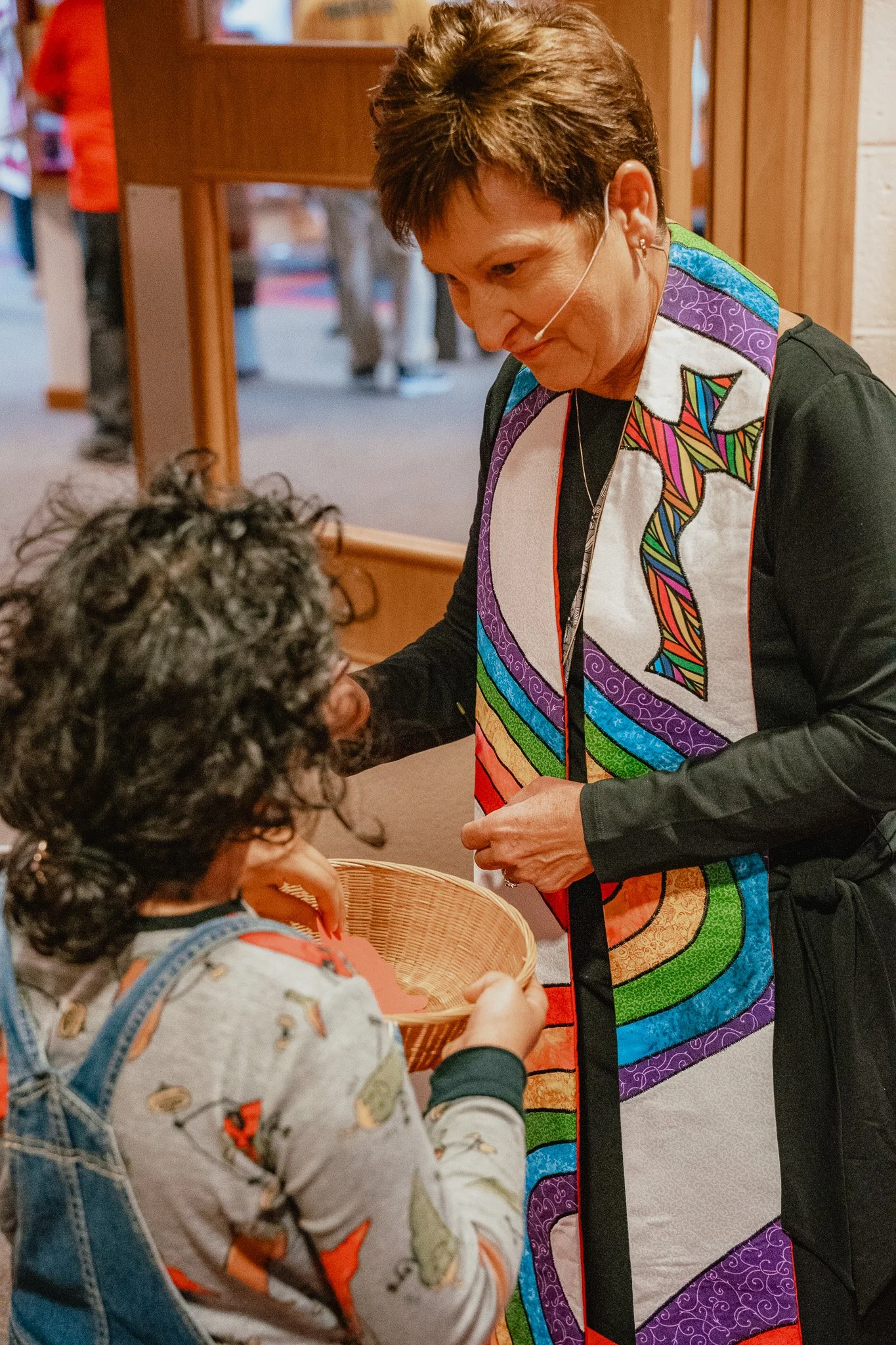A woman in a colorful, rainbow-striped stained glass pattern stole and apron hands a basket to a young girl with curly hair, wearing a floral shirt and denim overalls, inside of a wooden building.