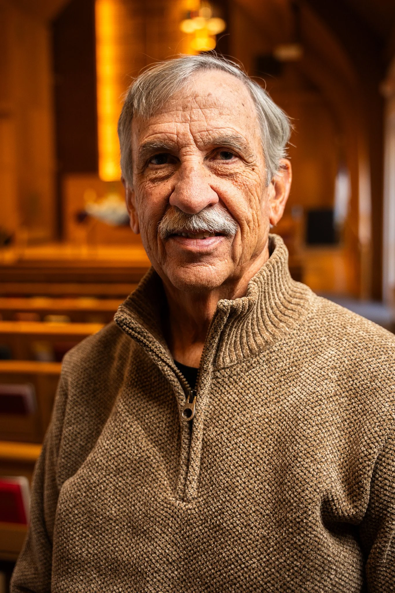 An elderly man with gray hair, a mustache, and a warm smile, wearing a brown knit sweater, standing inside a church with wooden furnishings.