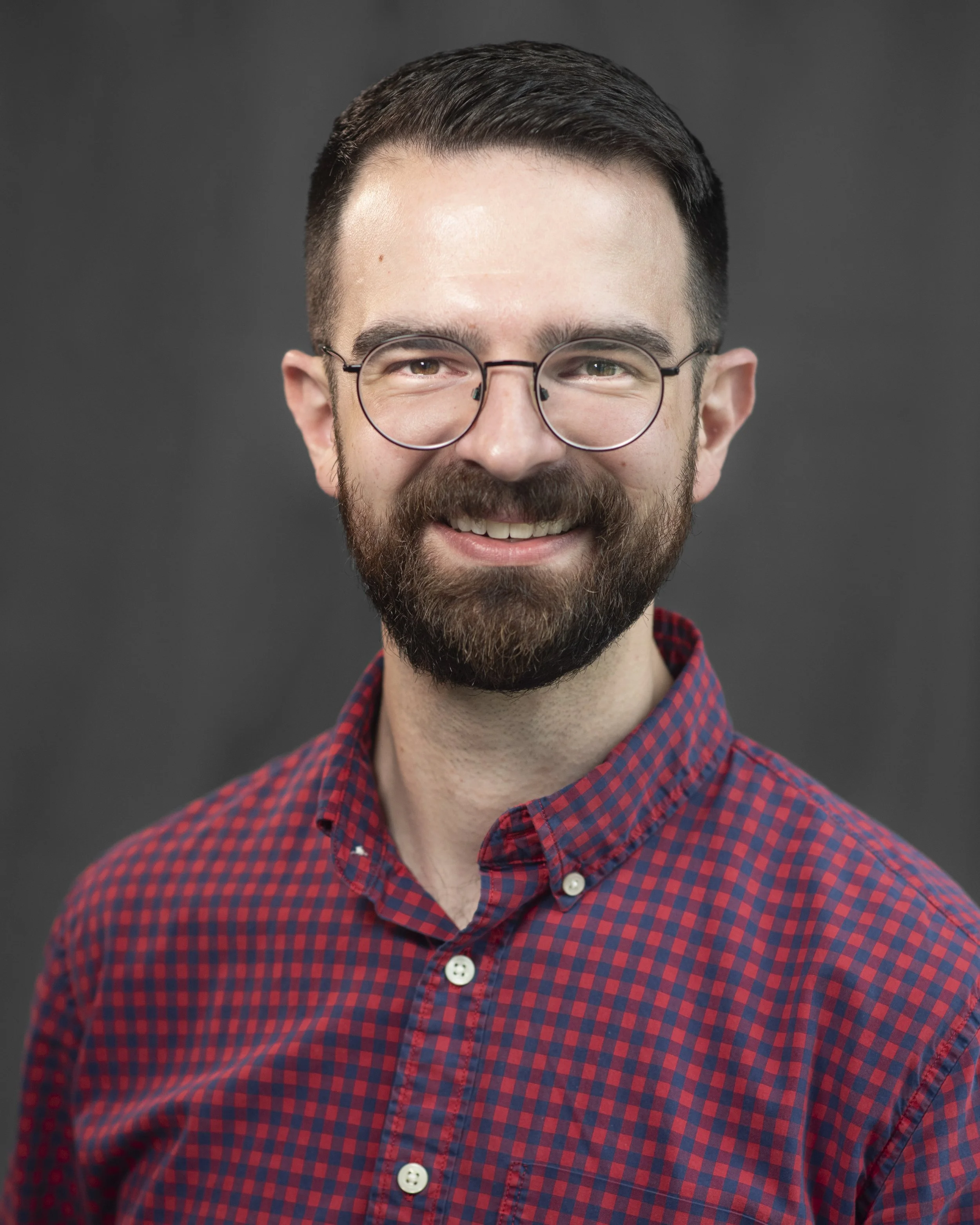 A smiling man with dark hair, glasses, and a beard wearing a red and blue checkered shirt standing inside a warmly lit church.