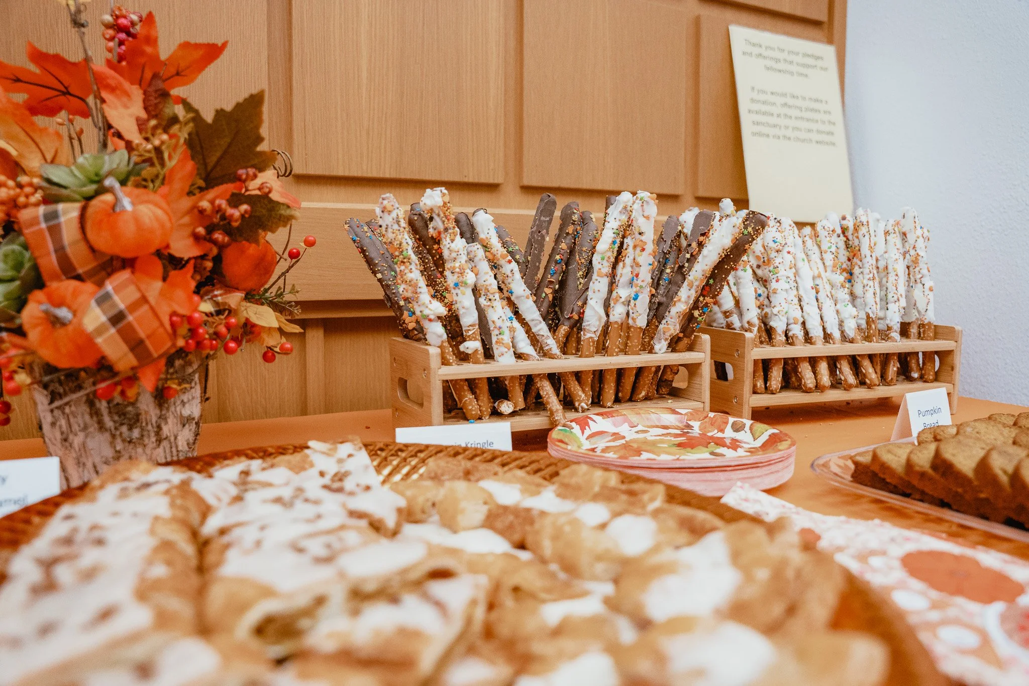A table display of Halloween-themed baked goods, featuring pretzel rods covered in white chocolate and festive sprinkles, along with other desserts like cookies and bread, with an autumn floral arrangement to the side.