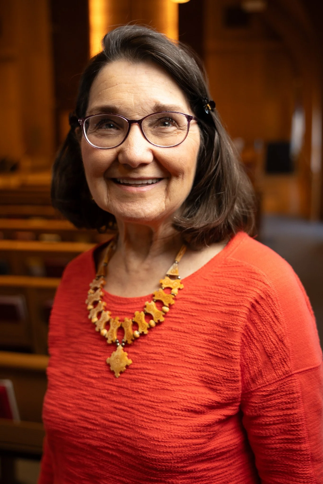 A smiling woman with shoulder-length brown hair, wearing glasses, a red textured top, and an intricate gold necklace, standing inside a wood-paneled room with chairs in the background.