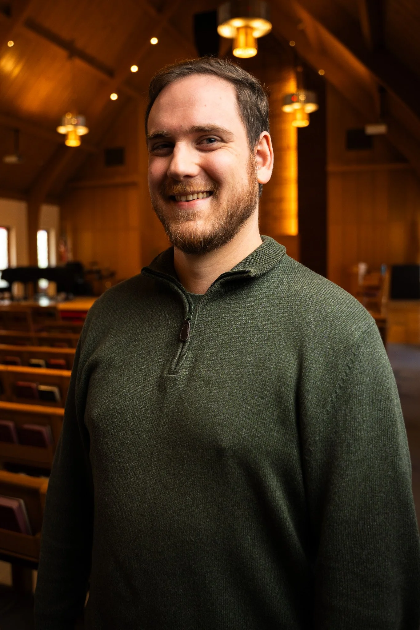 A smiling man with a beard and short hair standing inside a wooden church or hall, wearing a green sweater with a zip collar.